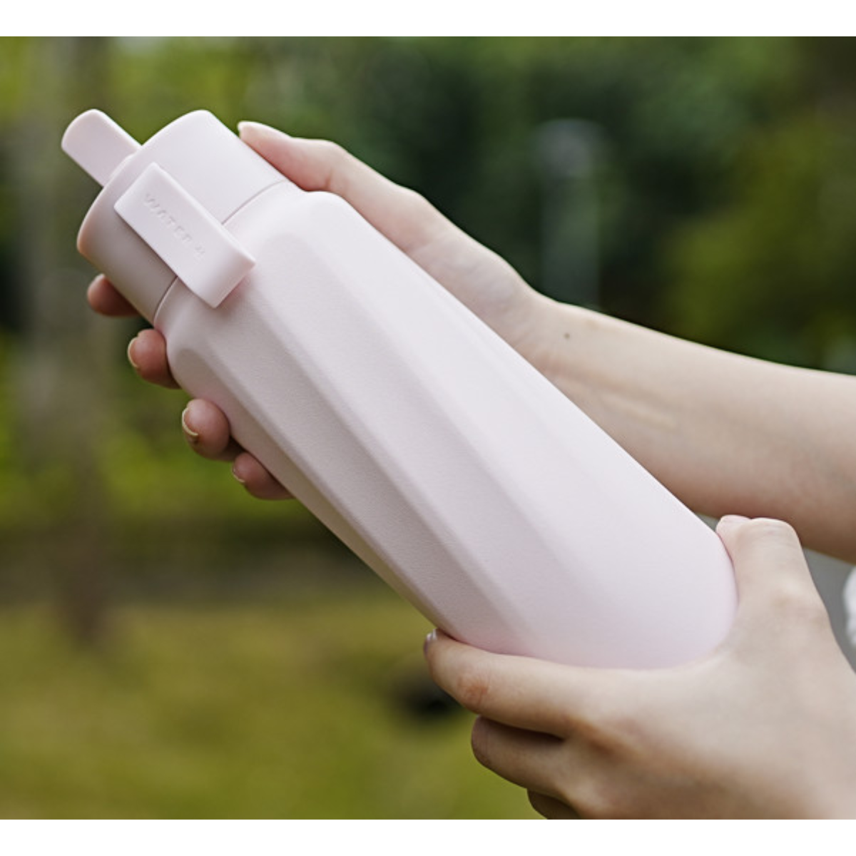 Eine Person hält eine matte, rosa Wasserflasche mit facettiertem Design vor einem verschwommenen grünen Hintergrund.