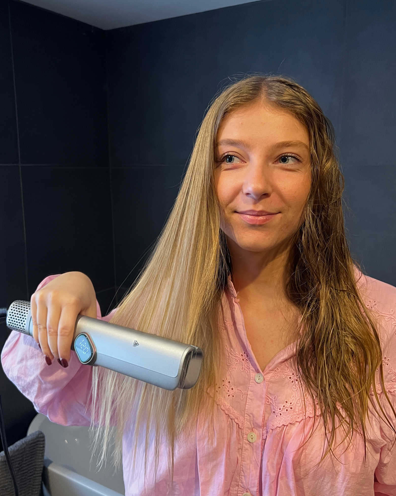 Una donna bionda con una camicia rosa liscia un lato dei capelli con uno styler argentato.