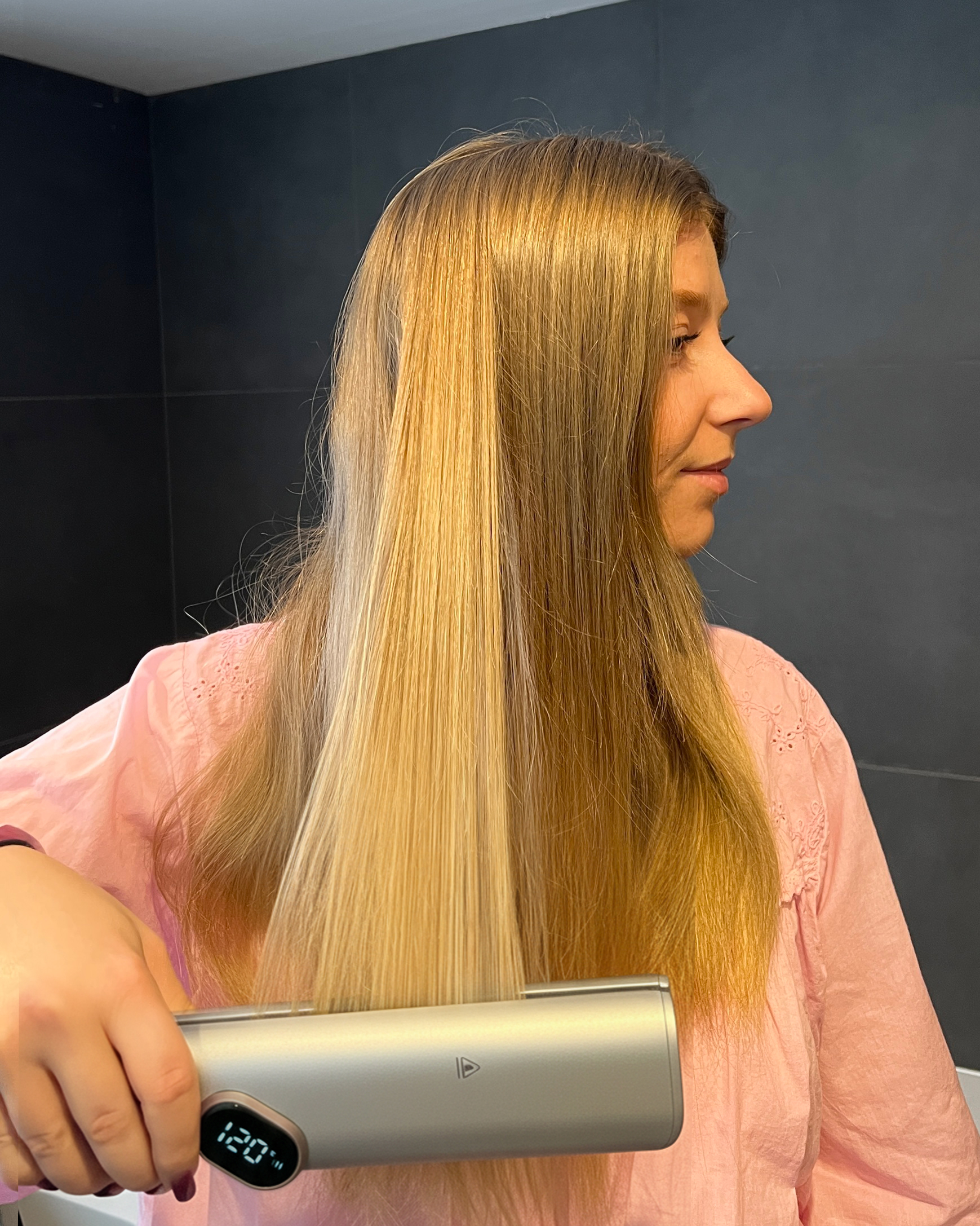 Una donna bionda liscia i suoi lunghi capelli con una piastra argentata in una stanza con piastrelle scure.