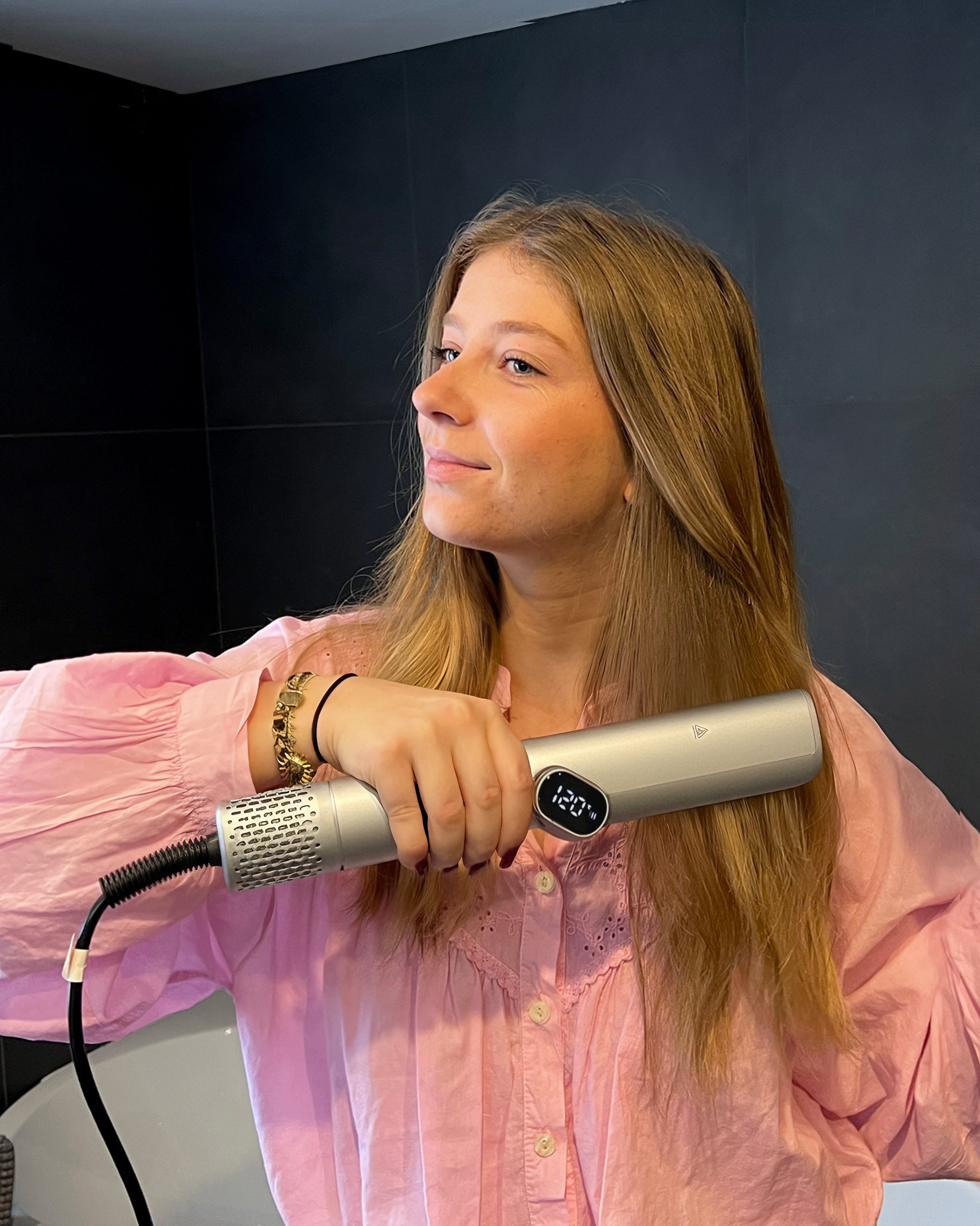 Una donna in camicia rosa tiene una piastra per capelli argentata, acconciando i capelli biondi su uno sfondo scuro.