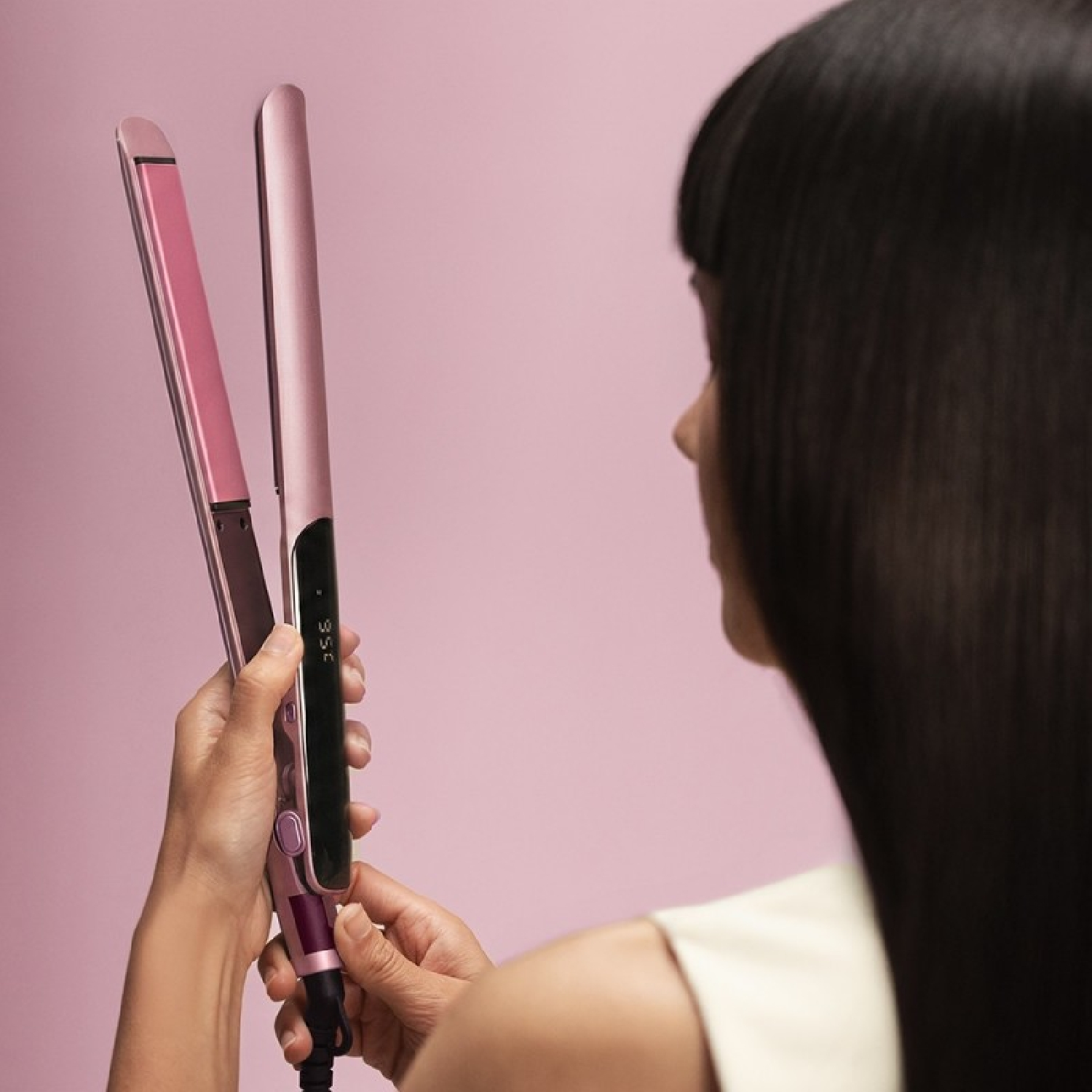Una donna con lunghi capelli scuri tiene una piastra per capelli rosa su sfondo rosa.