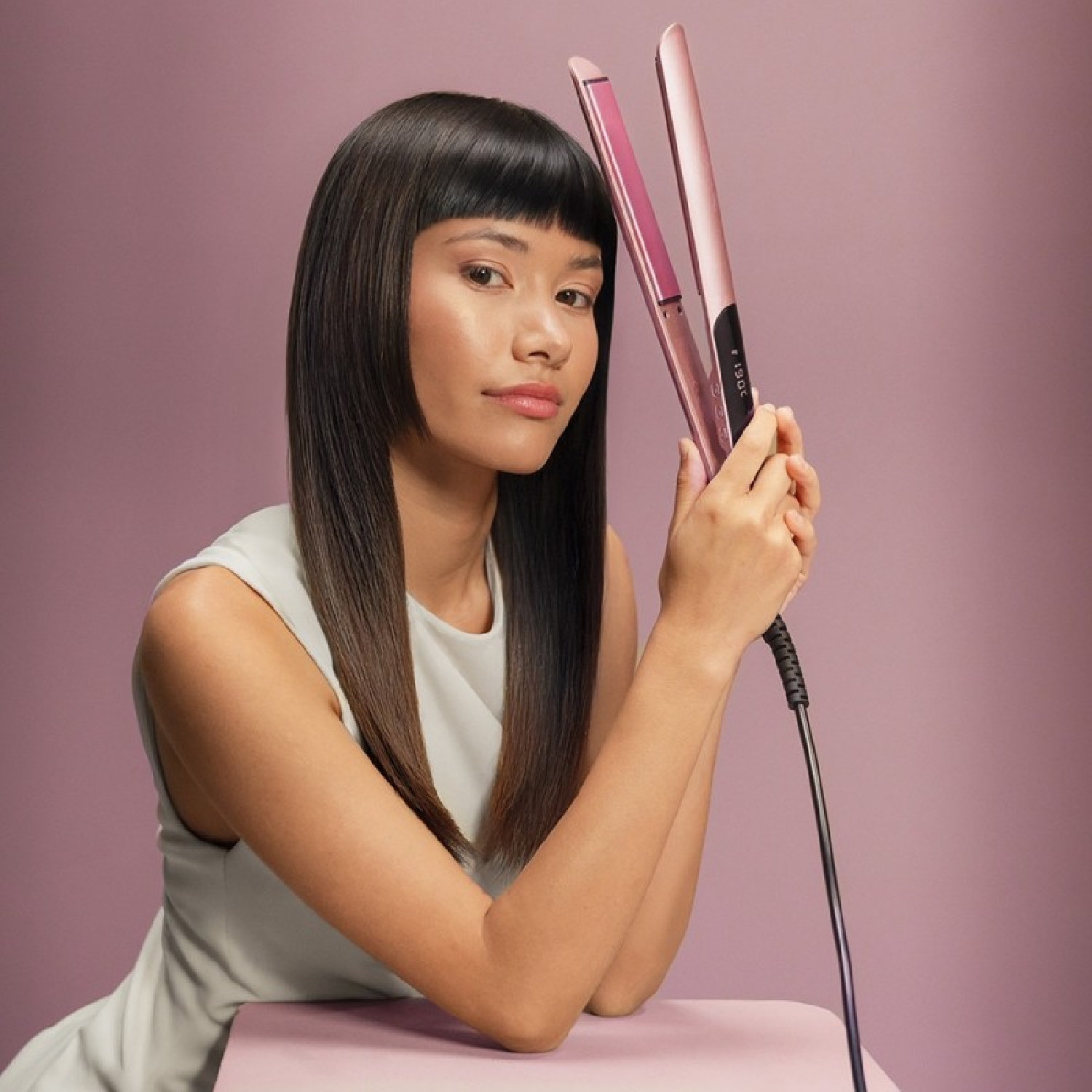 Una donna con capelli scuri e lisci tiene una piastra per capelli rosa su sfondo viola.