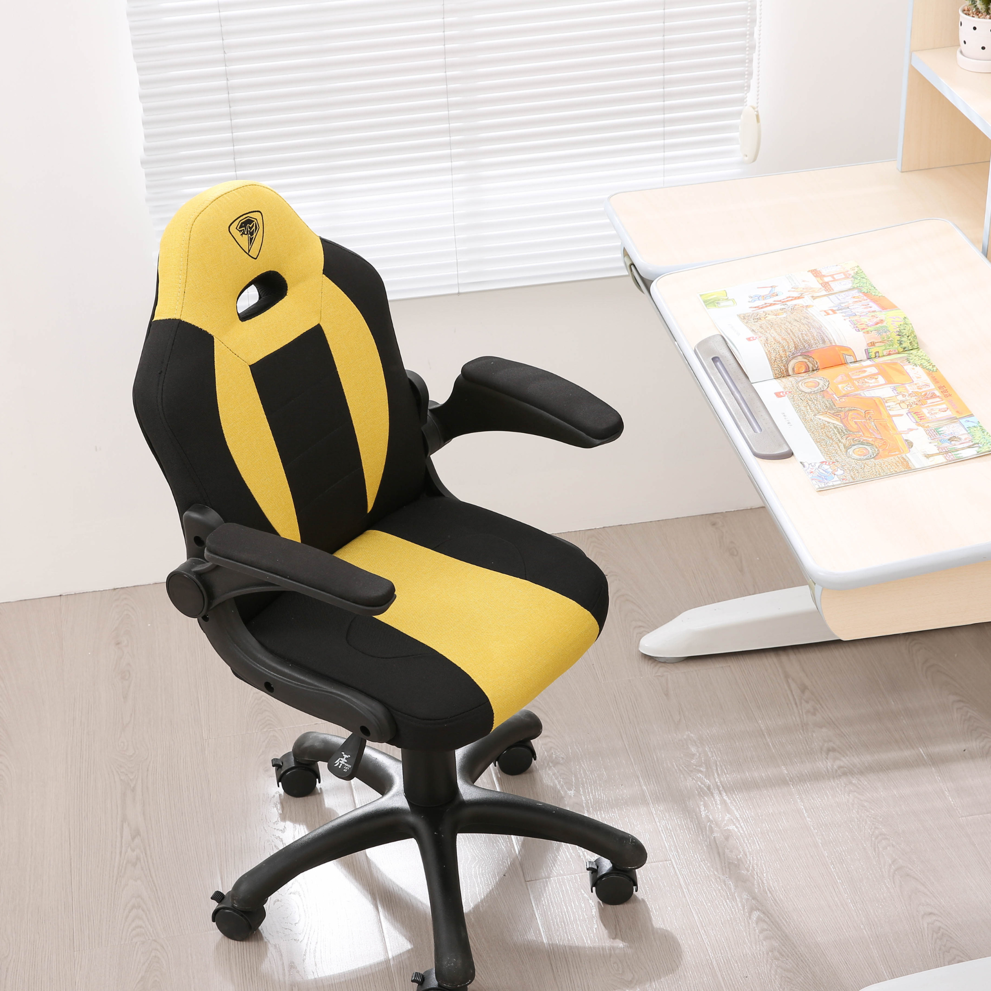Czarno-żółte ergonomiczne krzesło biurkowe stoi obok drewnianego biurka z otwartą książką w jasnym pokoju.