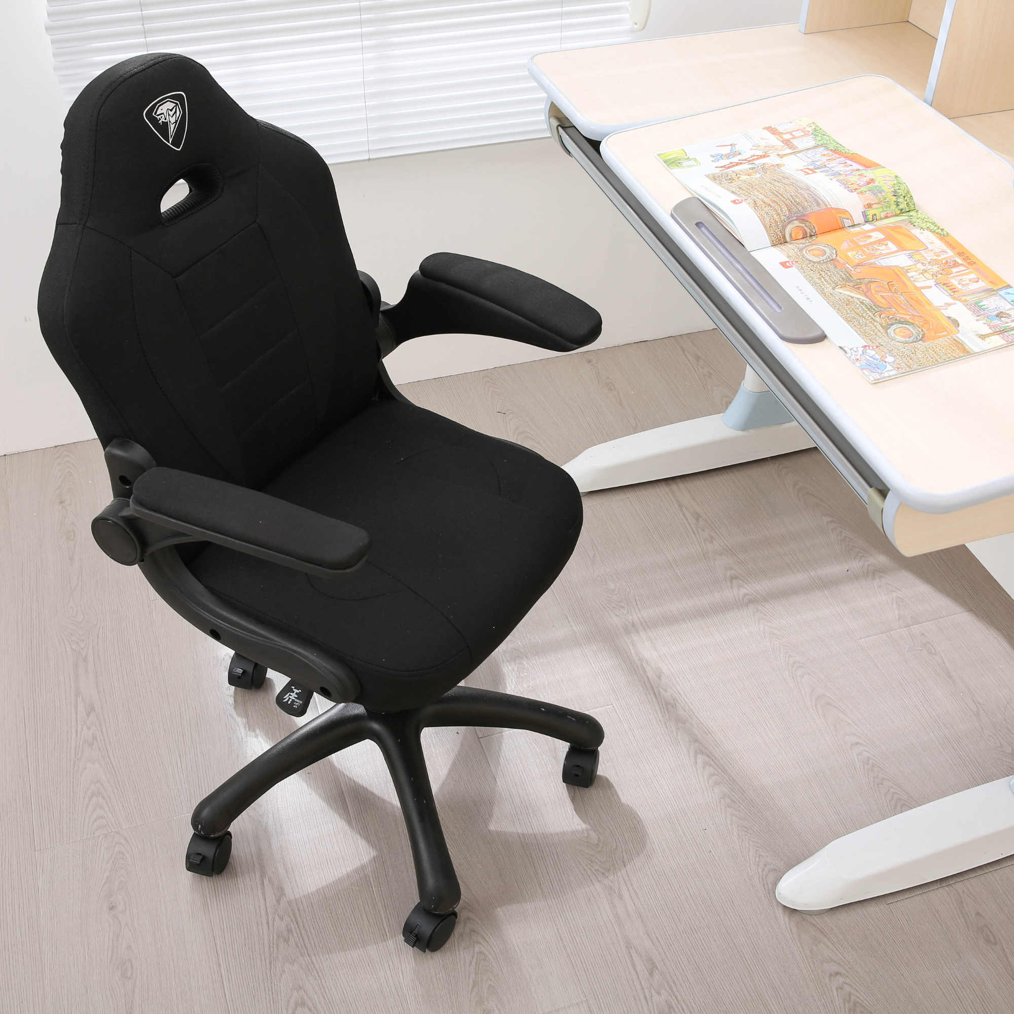 Czarne ergonomiczne krzesło biurkowe stoi na jasnej drewnianej podłodze obok biurka z otwartą książką.