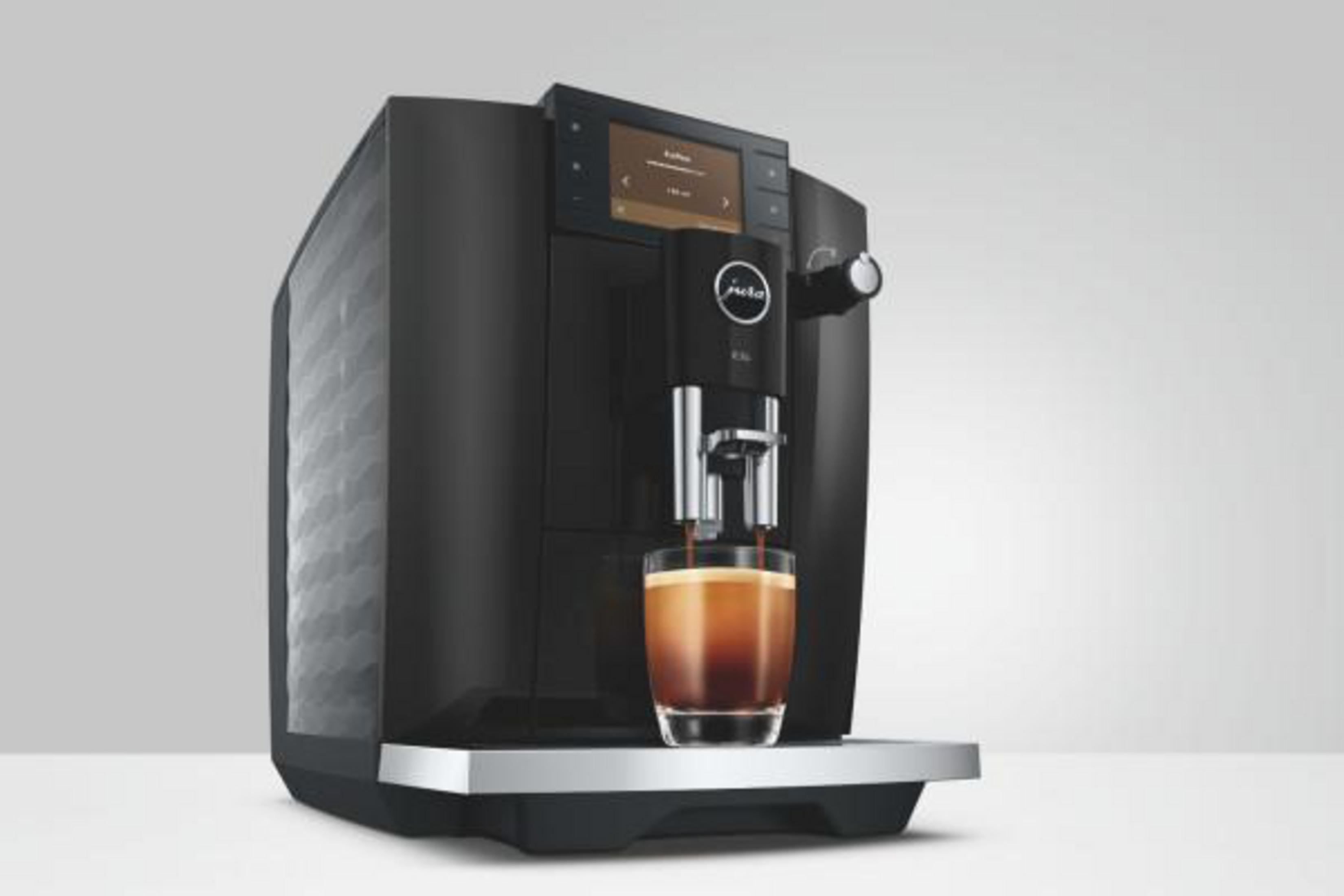 Una macchina da caffè Jura nera eroga caffè in un bicchiere di vetro con schiuma, su sfondo grigio chiaro.