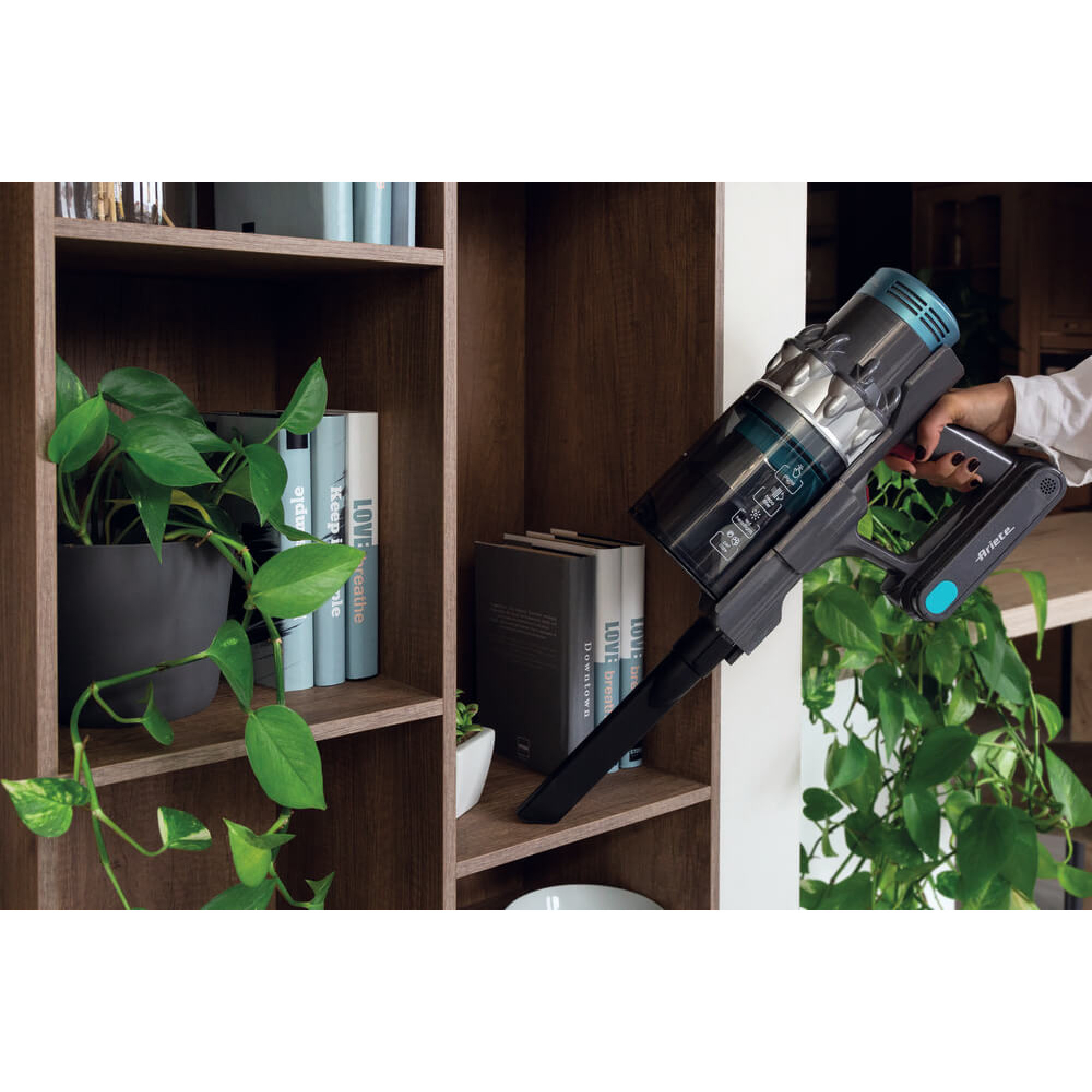 Aspirapolvere portatile tenuto davanti a una libreria in legno con libri e piante.