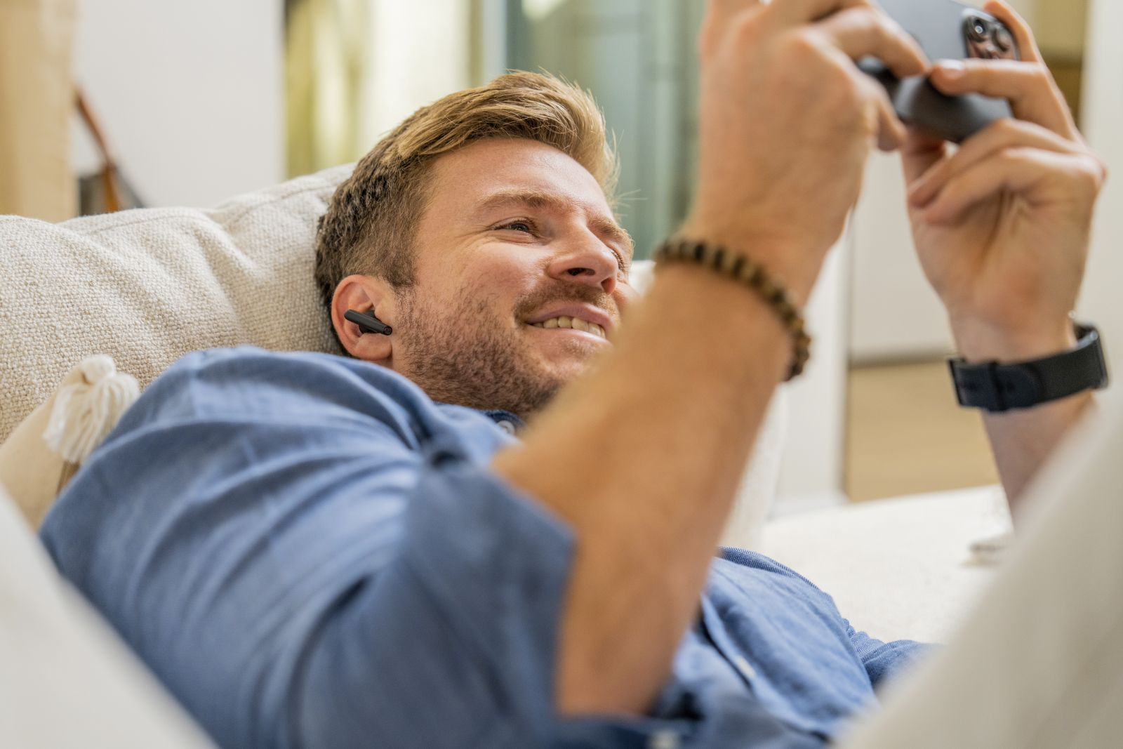Un homme blond allongé sur un canapé, portant des écouteurs intra-auriculaires et utilisant un smartphone.