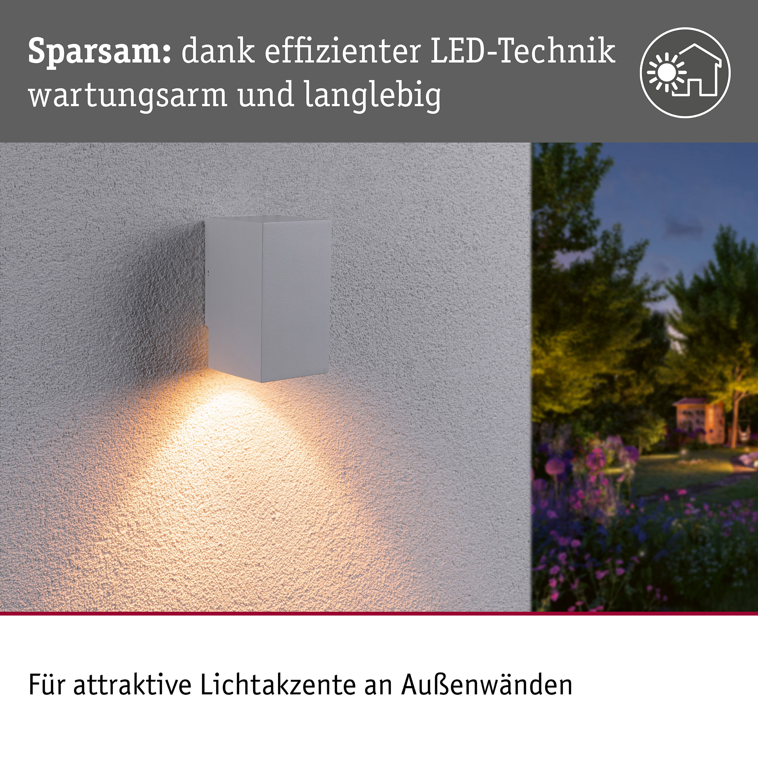Eine graue Wandleuchte wirft warmes Licht nach unten an eine Außenwand, im Hintergrund ein Garten.