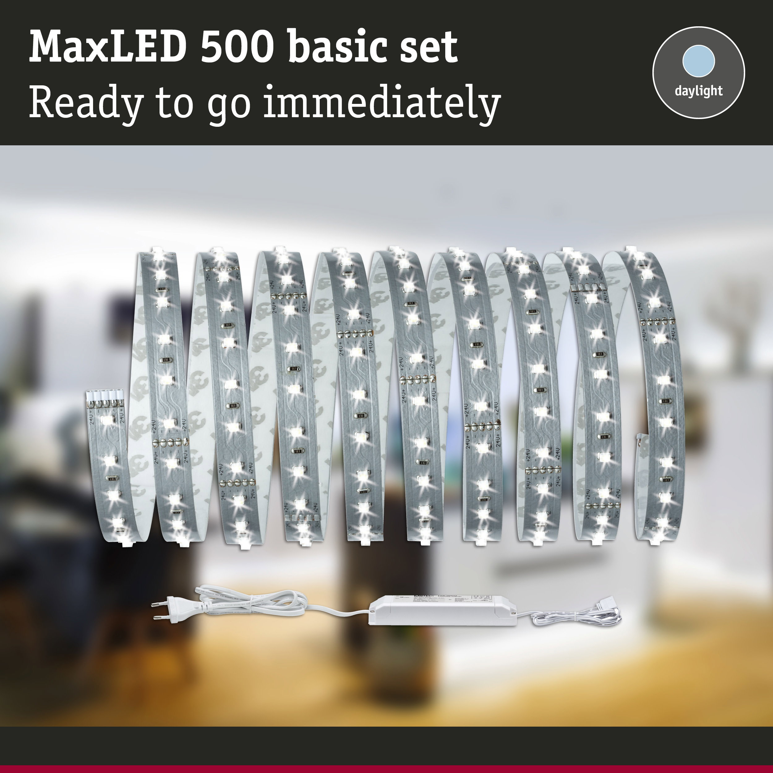 MaxLED 500 Basisset mit gerolltem, beleuchtetem LED-Streifen, Netzteil und Stecker vor verschwommenem Zimmerhintergrund.