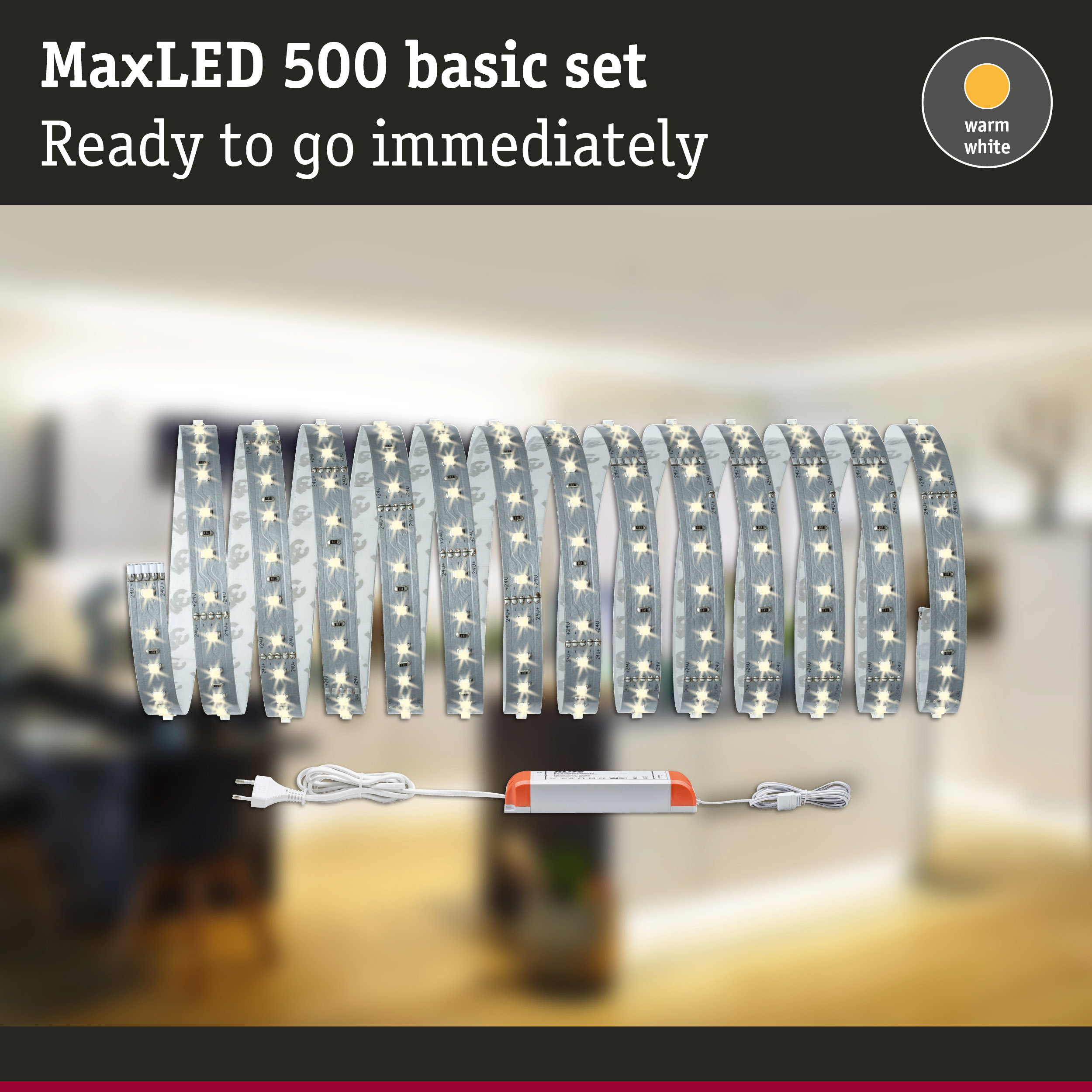 Set base MaxLED 500 con striscia LED arrotolata orizzontalmente e alimentatore su sfondo sfocato.