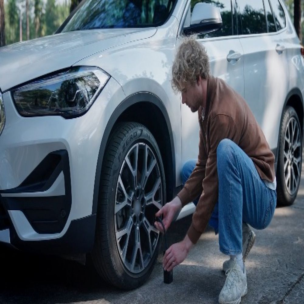 Ein blondgelockter Mann in Jeans und brauner Jacke pumpt den Reifen seines weißen Autos auf.