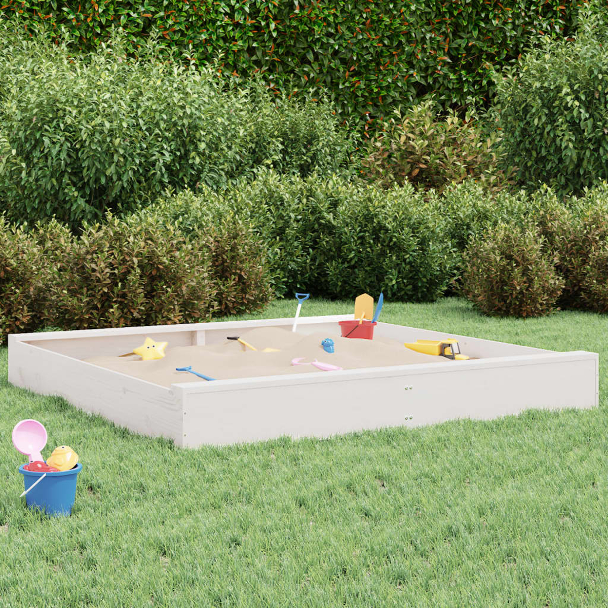 Ein weißer Holzsandkasten mit Sand und bunten Spielsachen steht auf einer Rasenfläche vor grünen Büschen.