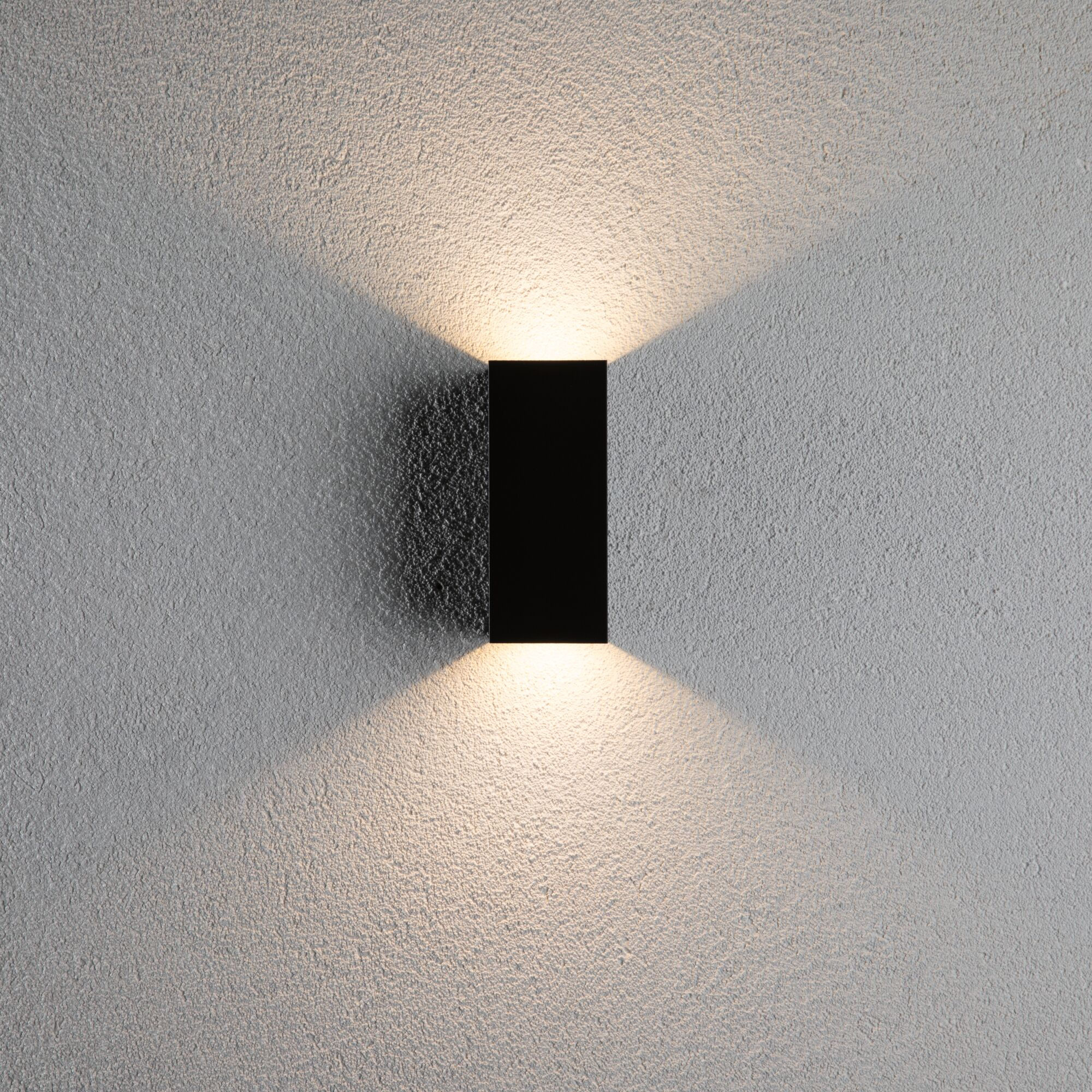 Czarna lampa ścienna rzuca stożki światła w górę i w dół na fakturowaną szarą ścianę.