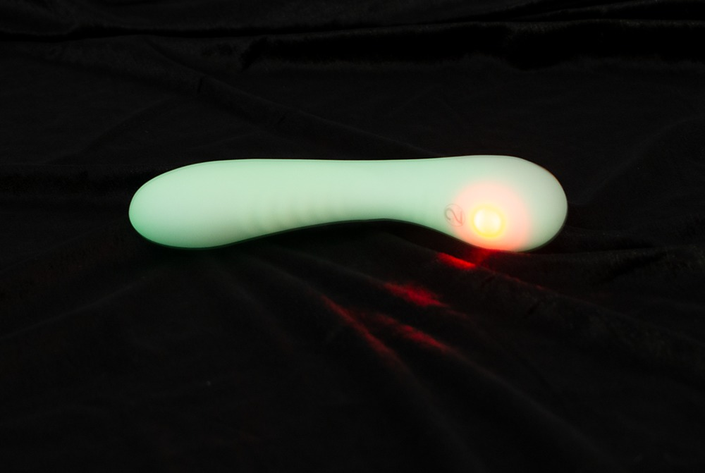 Ein grün leuchtendes, geschwungenes Wellness-Gerät mit einer orangefarbenen LED-Taste auf schwarzem Samthintergrund.