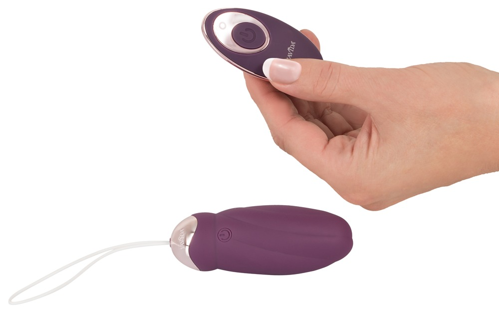 Eine Hand hält eine kleine, ovale, violette Fernbedienung mit kupferfarbenem Rand. Darunter liegt ein vibratorähnliches Gerät mit weißer Kordel vor weißem Hintergrund.
