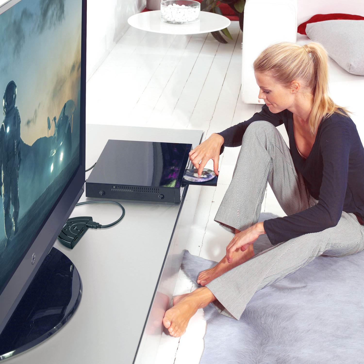 Una mujer rubia está sentada en el suelo insertando un DVD en un reproductor.
