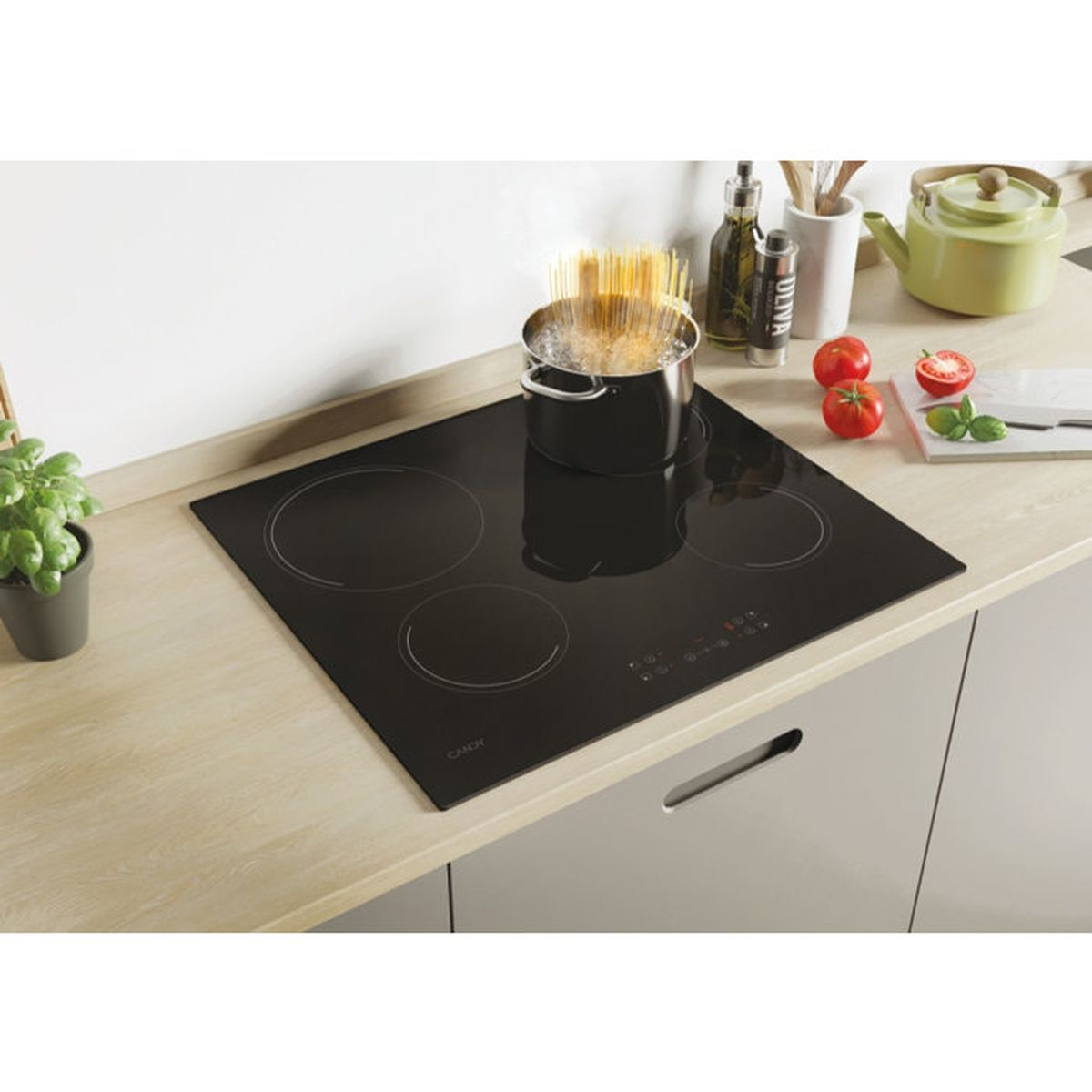 Una placa de inducción negra en encimera de madera, con olla de espaguetis hirviendo, tomates y tetera verde.