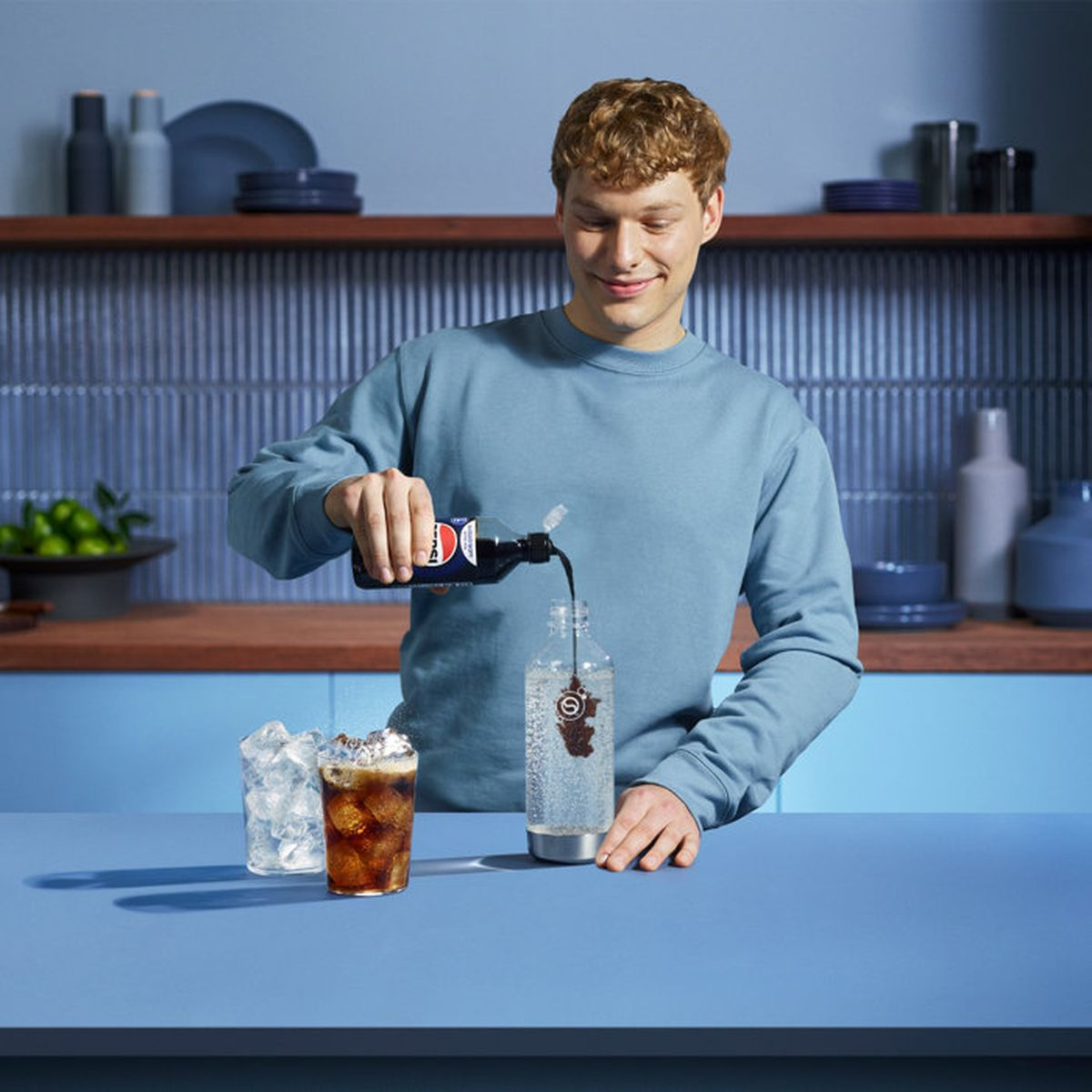 Ein Mann gießt Sirup in eine Wasserflasche auf einer blauen Theke mit Eisgläsern.