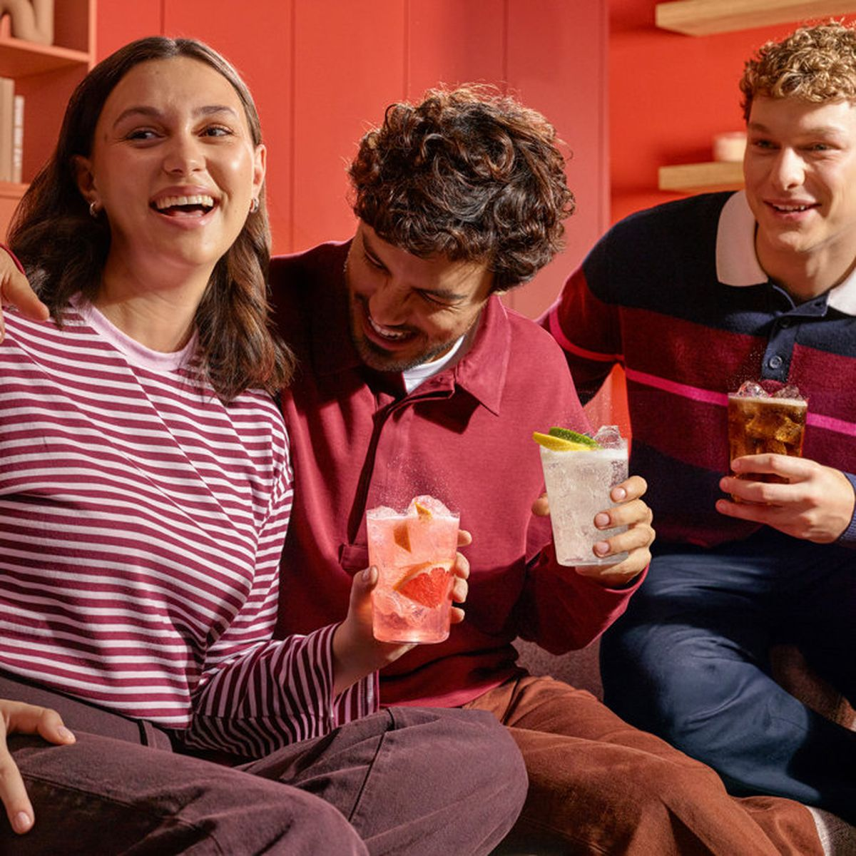 Tre giovani amici sorridono e tengono cocktail davanti a uno sfondo rosso all'interno.