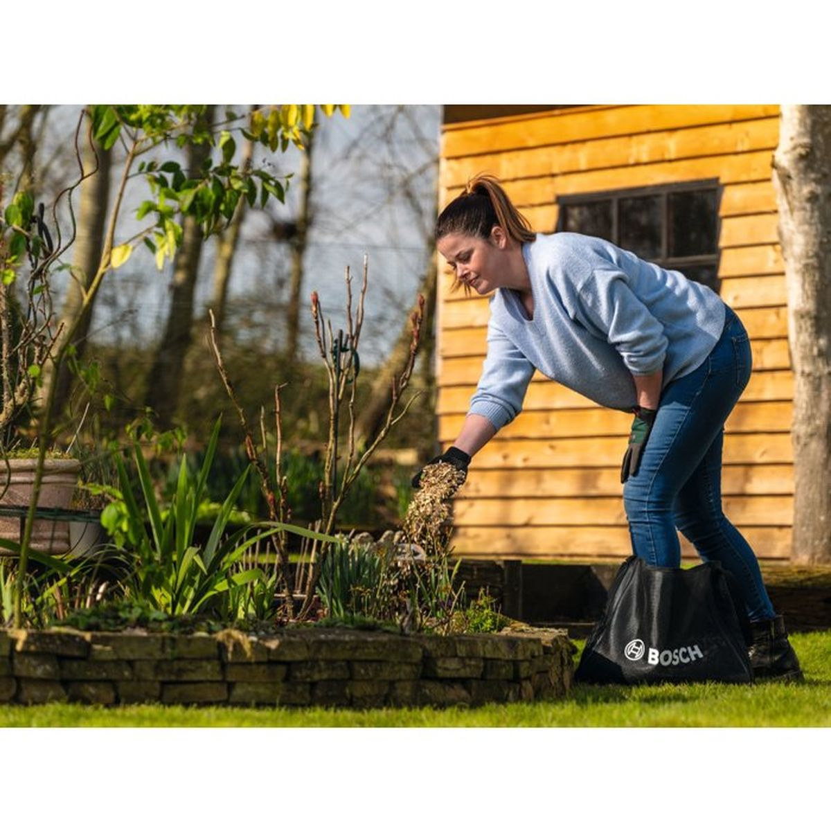 Eine Frau im blauen Pullover verteilt Mulch aus einer Bosch-Tasche in einem Gartenbeet vor einem Holzschuppen.