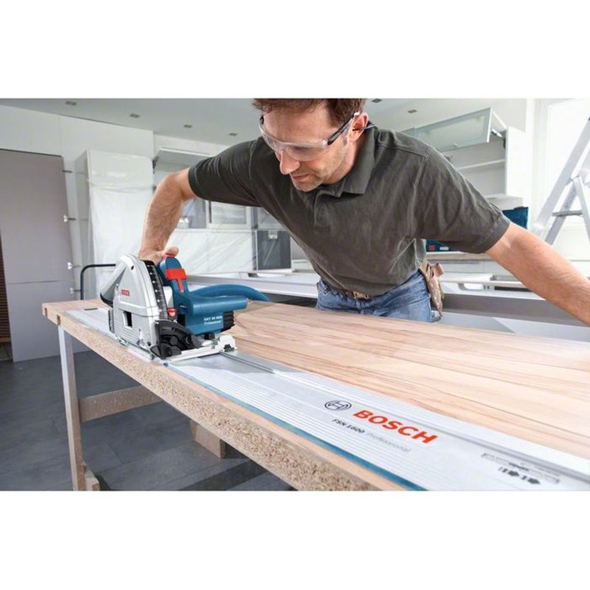 Ein Handwerker mit Schutzbrille schneidet eine Holzplatte mit einer blauen Bosch-Handkreissäge auf einer Schiene.