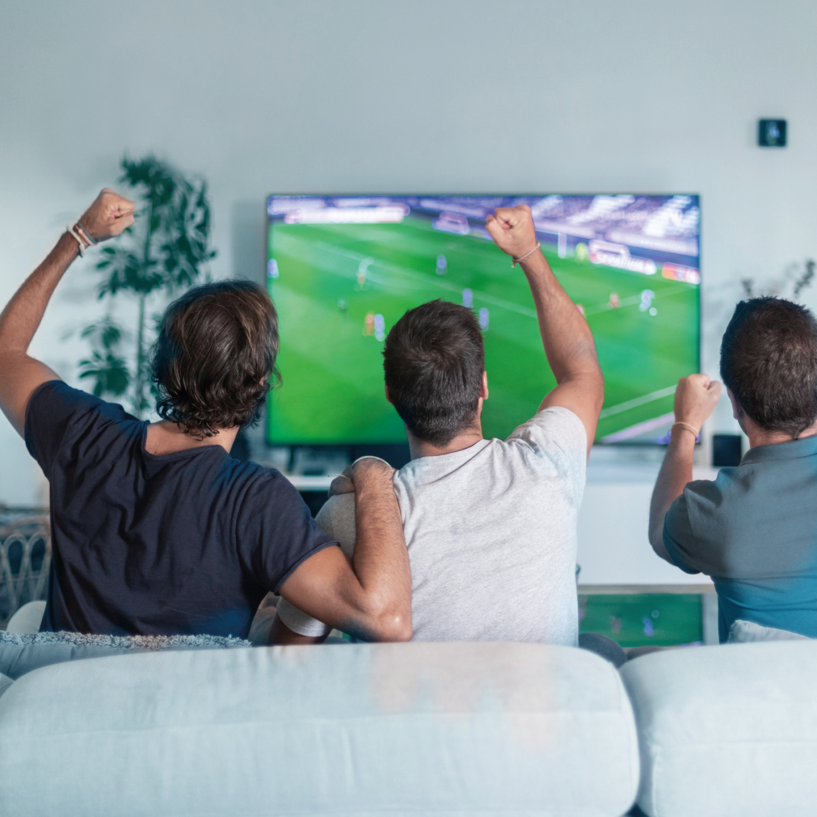 Tre uomini seduti su un divano esultano mentre guardano una partita di calcio su una grande TV.
