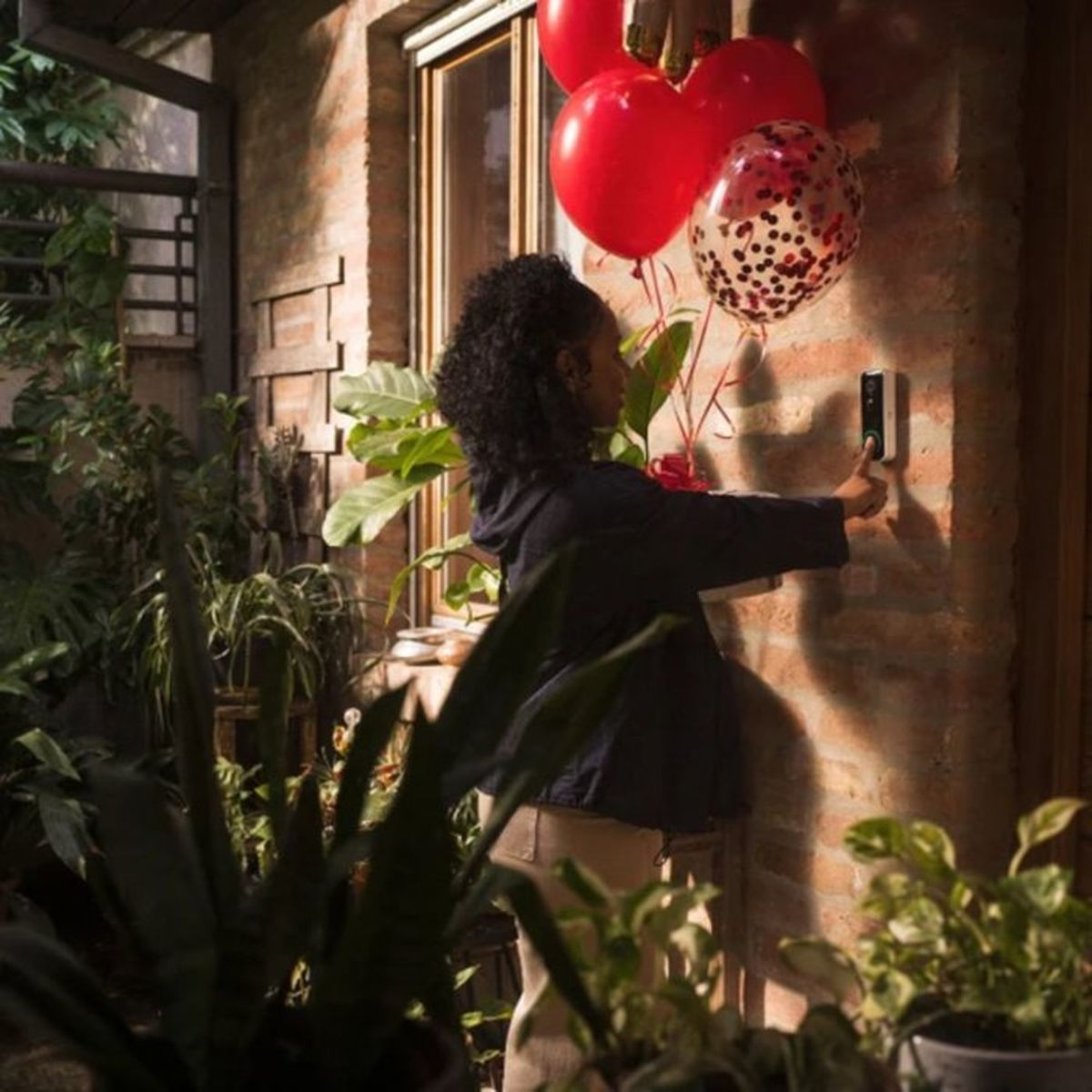 Una mujer con chaqueta negra presiona el timbre en una pared de ladrillo, rodeada de globos rojos y plantas.