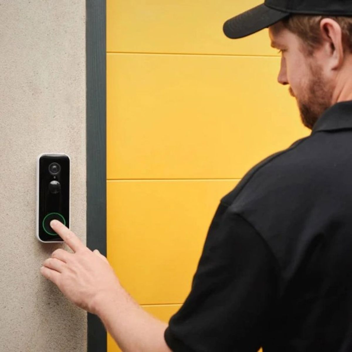 Un repartidor con ropa negra presiona el botón de un timbre con video en una pared junto a una puerta amarilla.