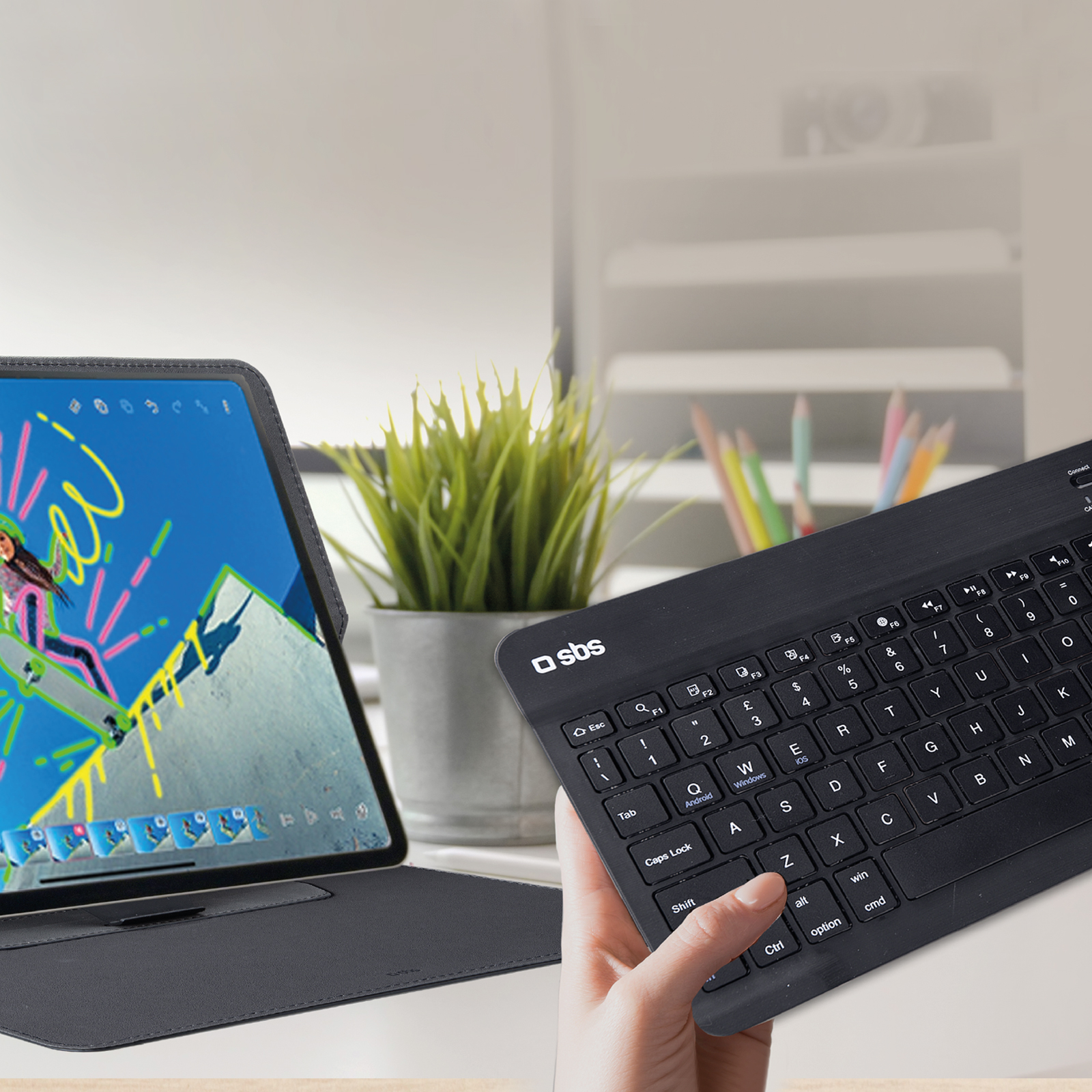 Mano sosteniendo un teclado negro junto a una tableta con gráficos y una planta en el fondo.