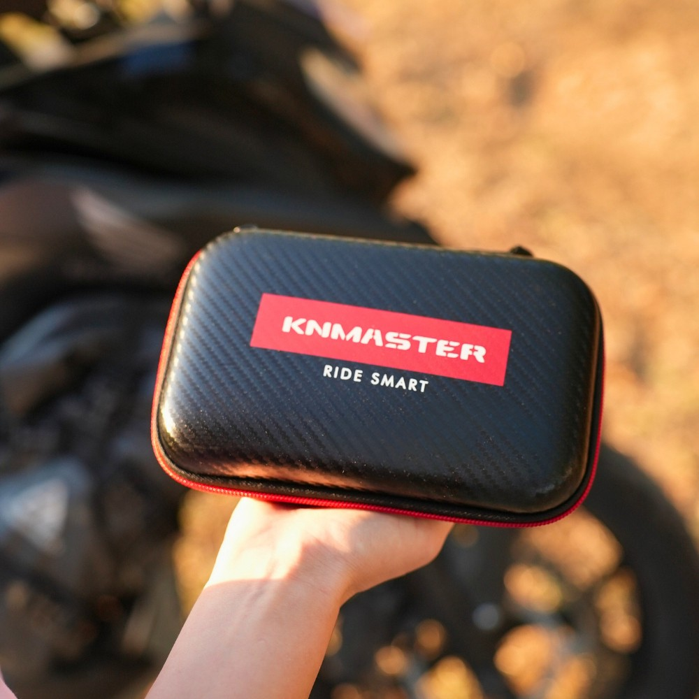 Bir el, bulanık bir motosikletin önünde kırmızı fermuarlı siyah bir KINMASTER sert çanta tutuyor.