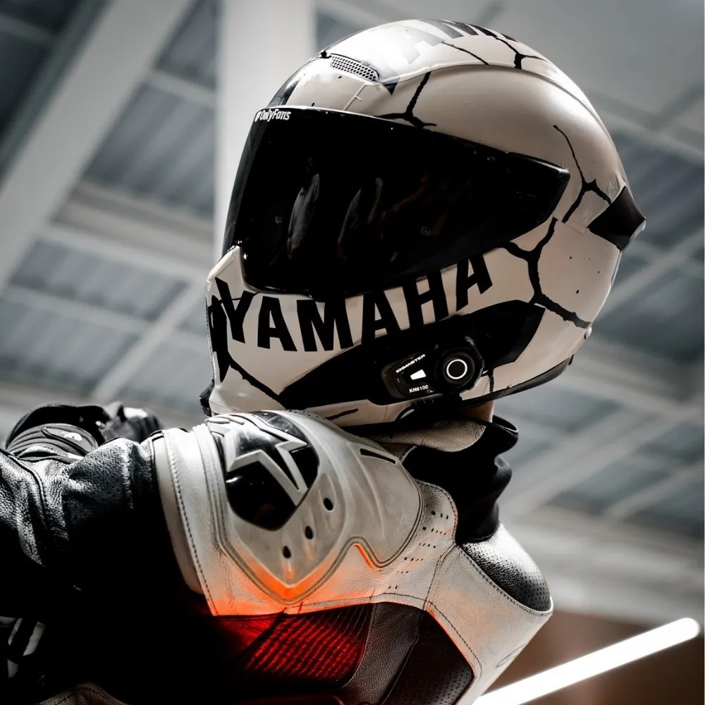 Garajda çatlak desenli beyaz Yamaha kaskı ve koruyucu giysileri olan bir motosiklet sürücüsü.