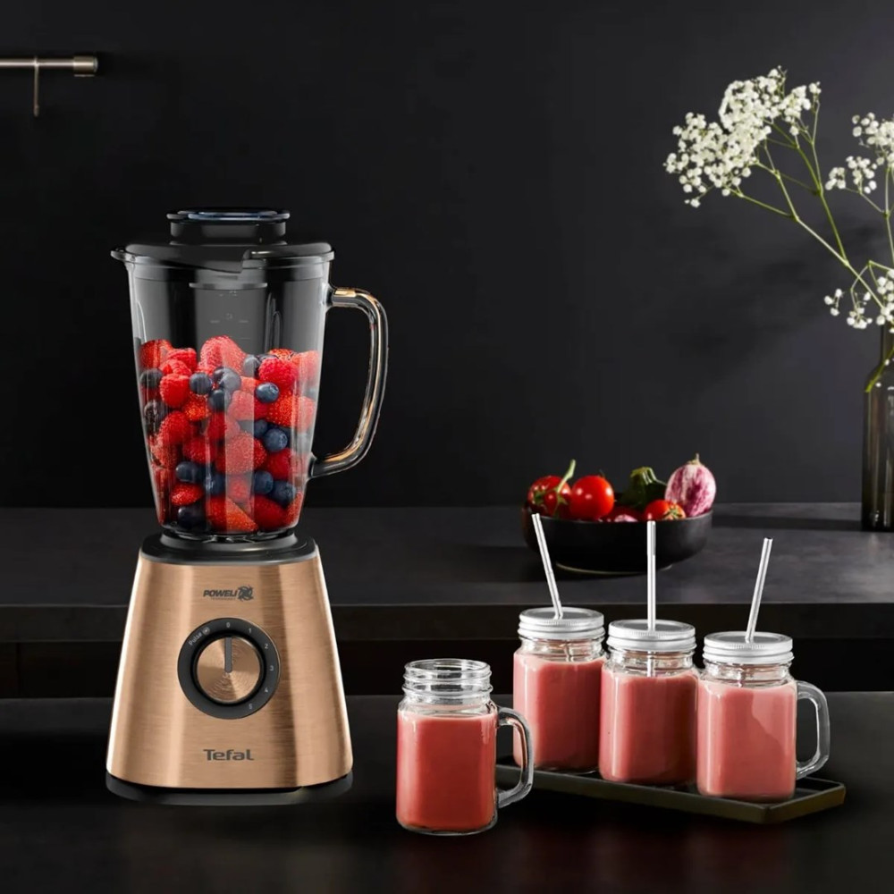 Złoty blender Tefal wypełniony owocami stoi obok czterech szklanek różowego smoothie na ciemnym blacie.