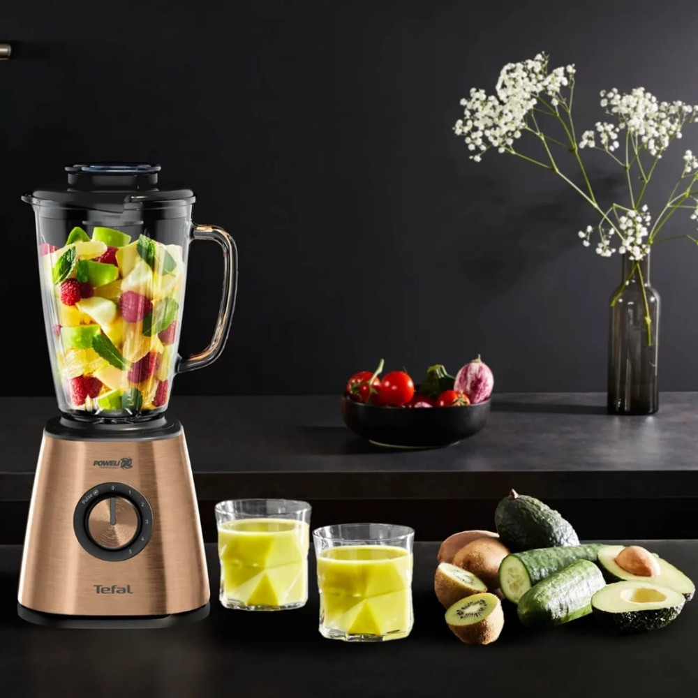 Różowozłoty blender Tefal z owocami, szklanki i awokado na czarnym stole, w tle kwiaty w wazonie.