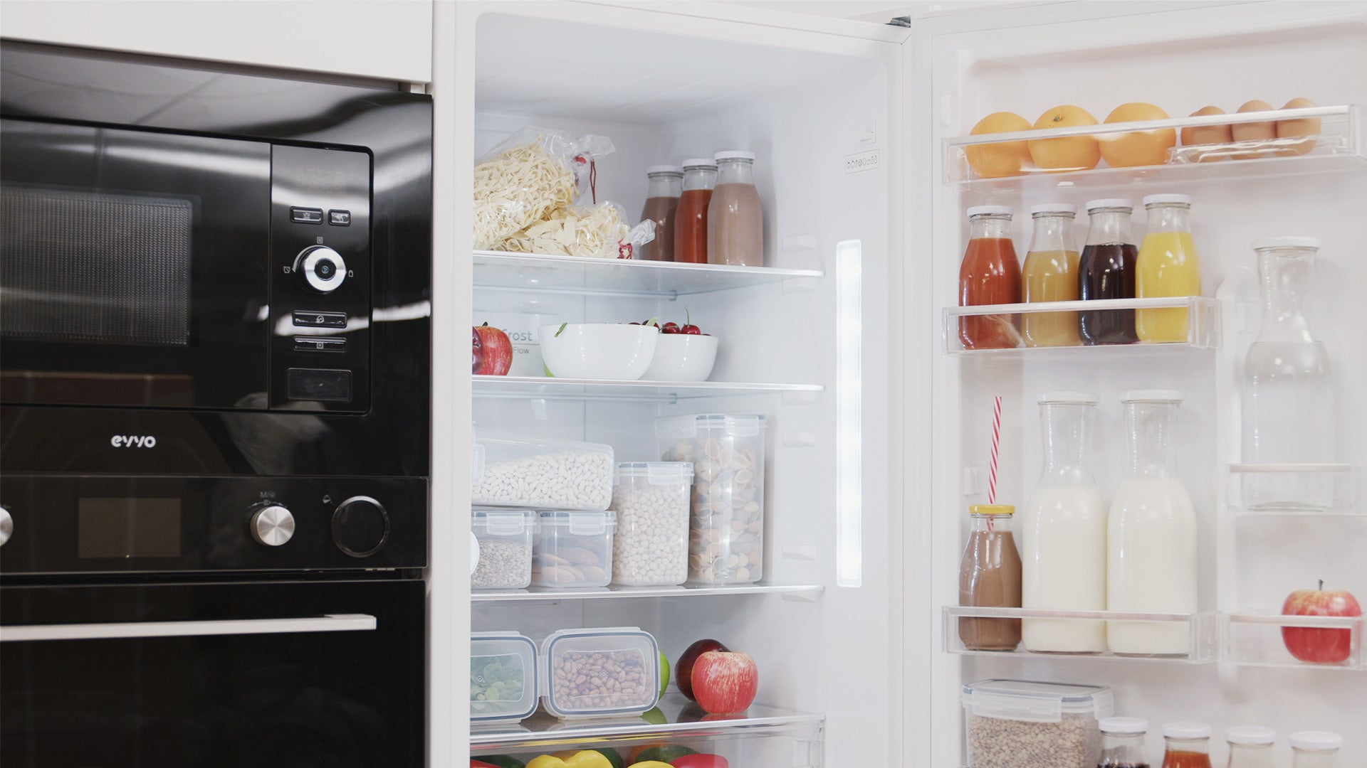 Un refrigerador blanco abierto lleno de comida, frascos y botellas junto a un horno empotrado negro.