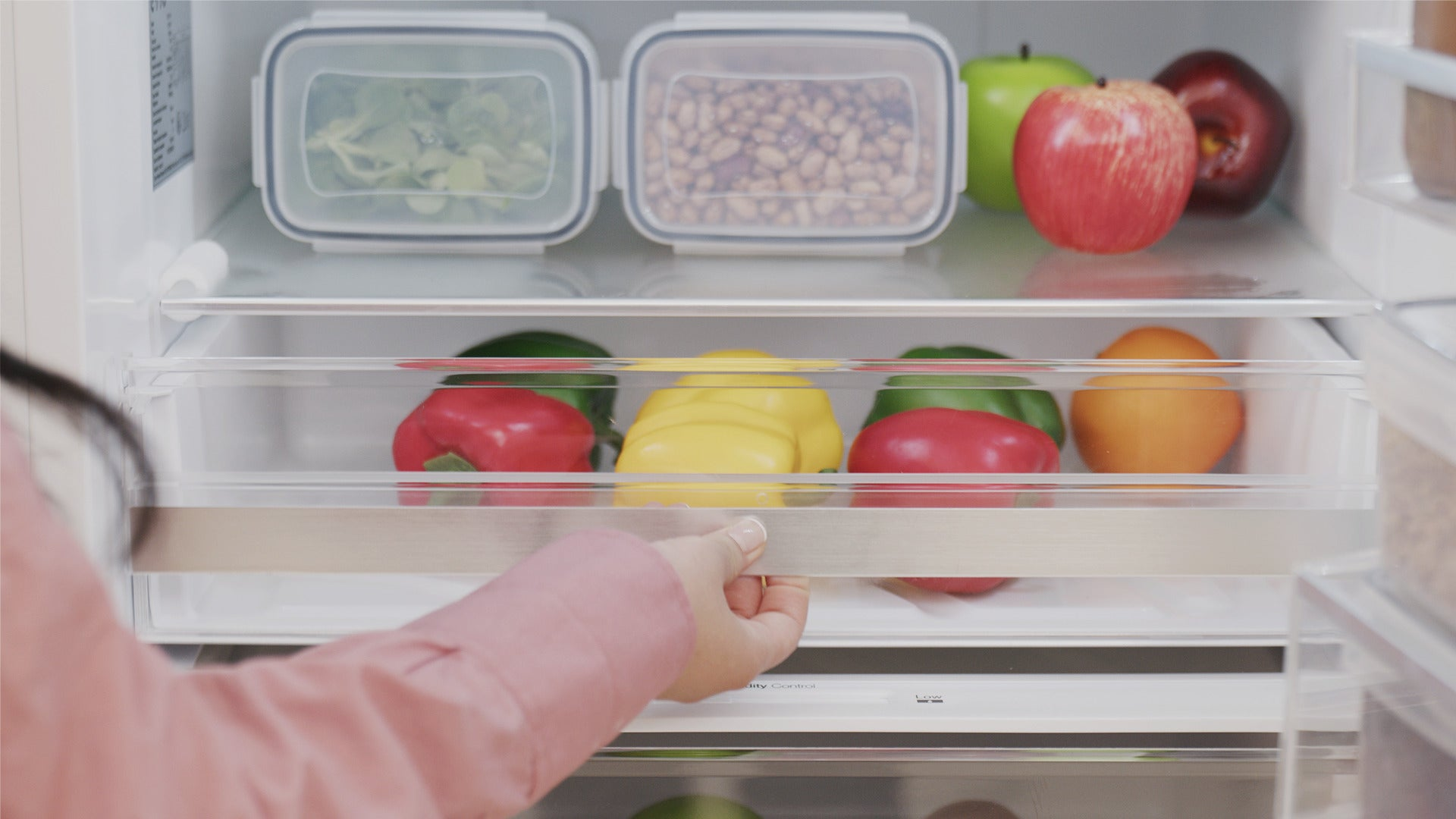 Una persona abre el cajón del refrigerador con pimientos de colores; arriba hay recipientes y manzanas.