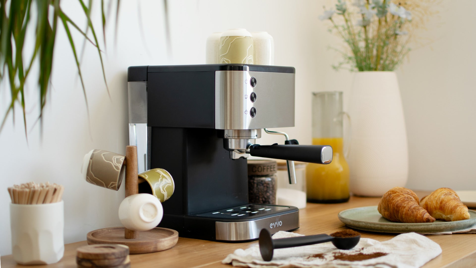 Una cafetera negra y plateada sobre una encimera de madera con cruasanes y tazas.
