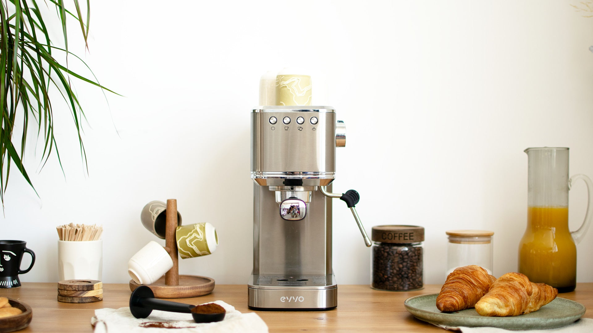 Una cafetera espresso de acero inoxidable en un mostrador de madera con cruasanes, granos de café, tazas y jarra.
