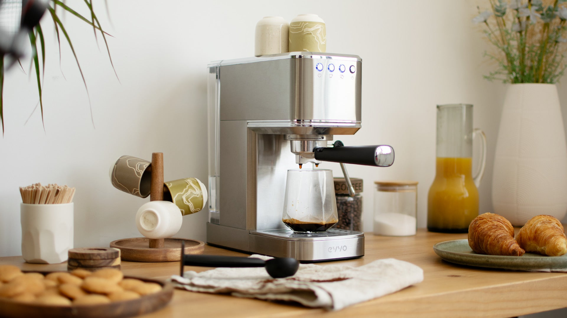 Una cafetera espresso de acero inoxidable en una barra de madera vierte café en un vaso, rodeada de cruasanes y tazas.