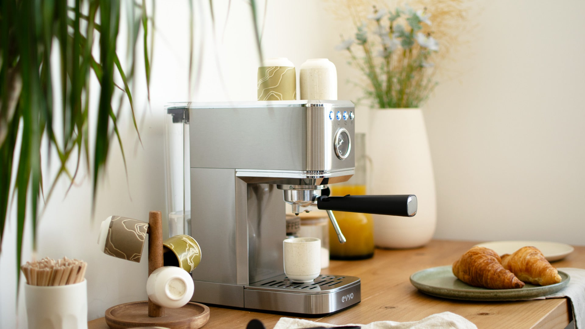 Una cafetera de espresso plateada en una barra de madera, junto a tazas pequeñas y dos cruasanes.