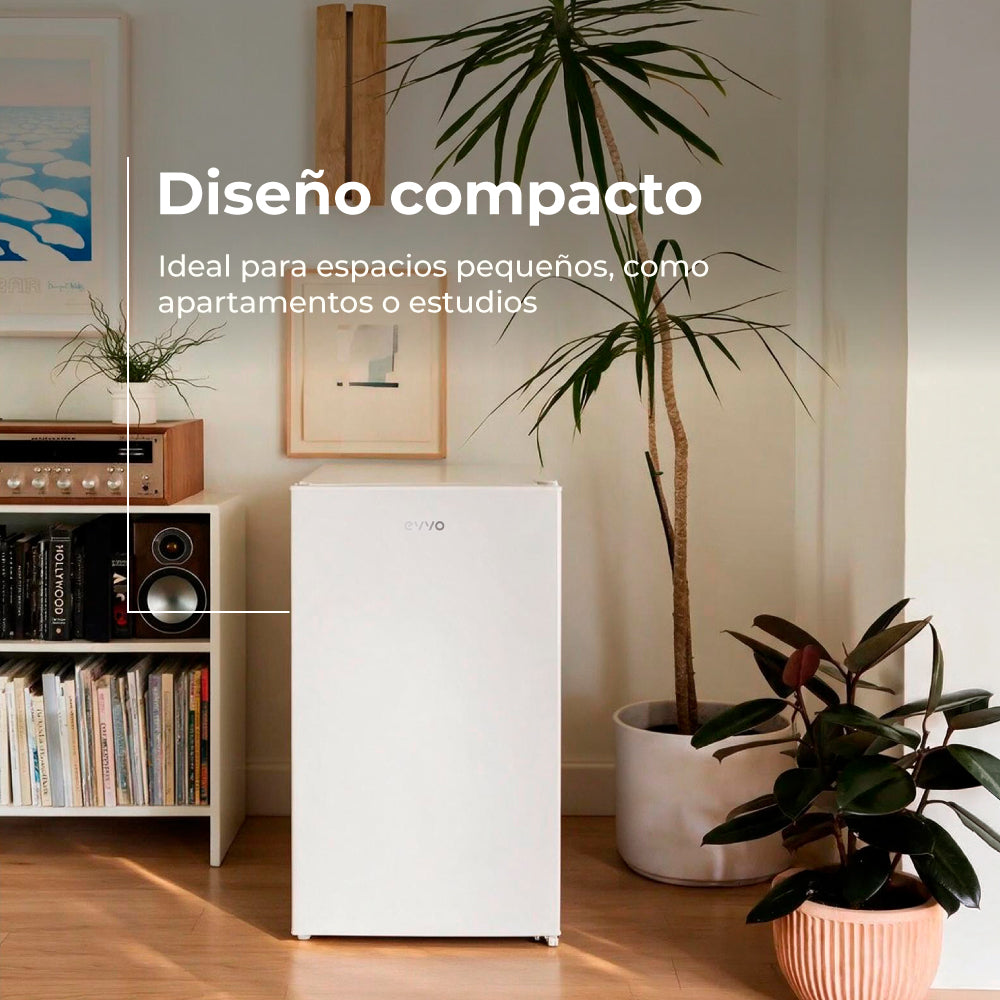 Una nevera blanca y compacta se encuentra en una habitación luminosa junto a plantas y una estantería.