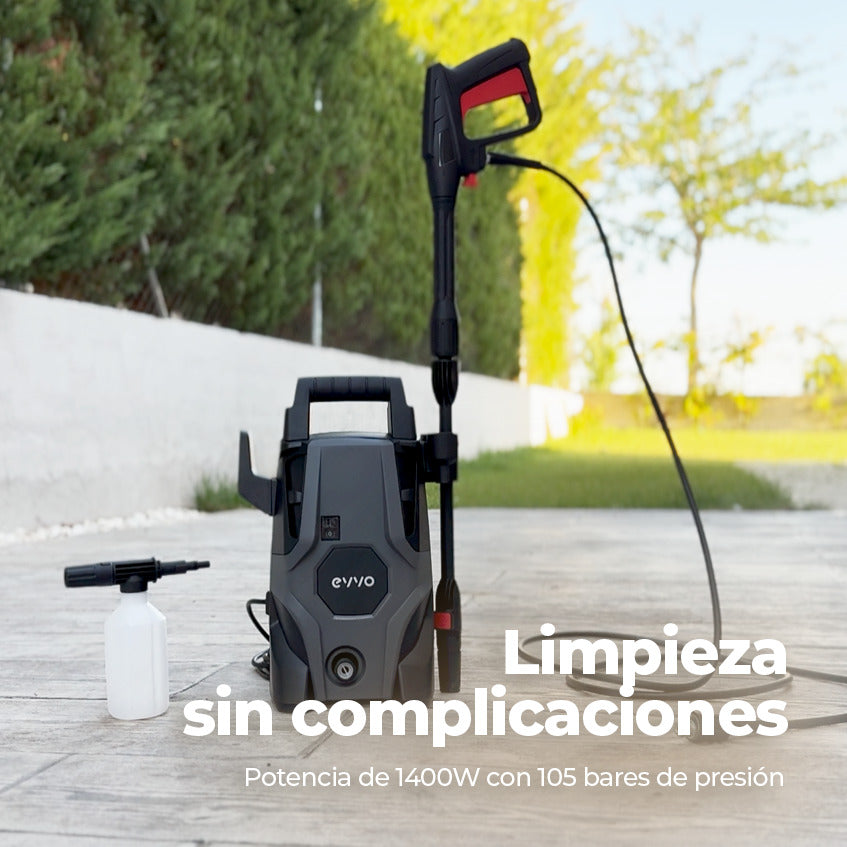 Hidrolimpiadora gris en una terraza con manguera y accesorios frente a un seto verde.