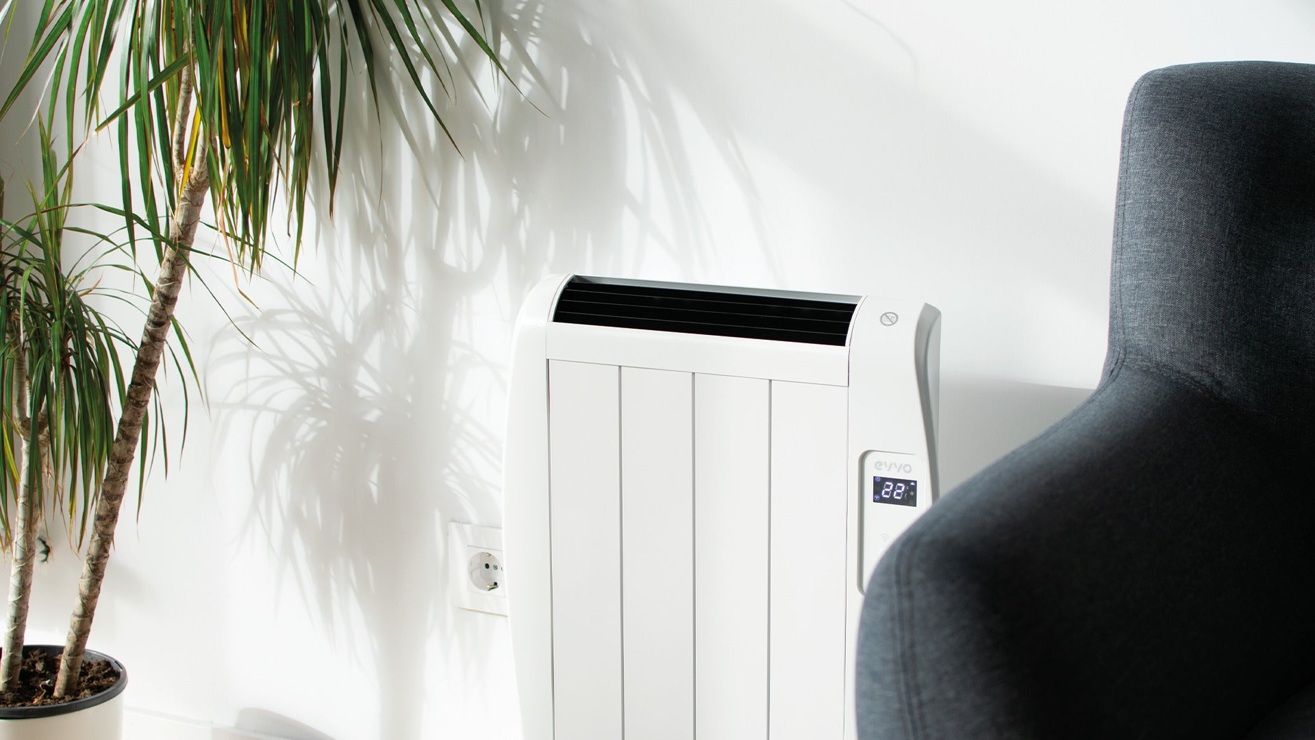 Un radiador blanco contra una pared, con una planta en maceta y un sillón oscuro cerca.