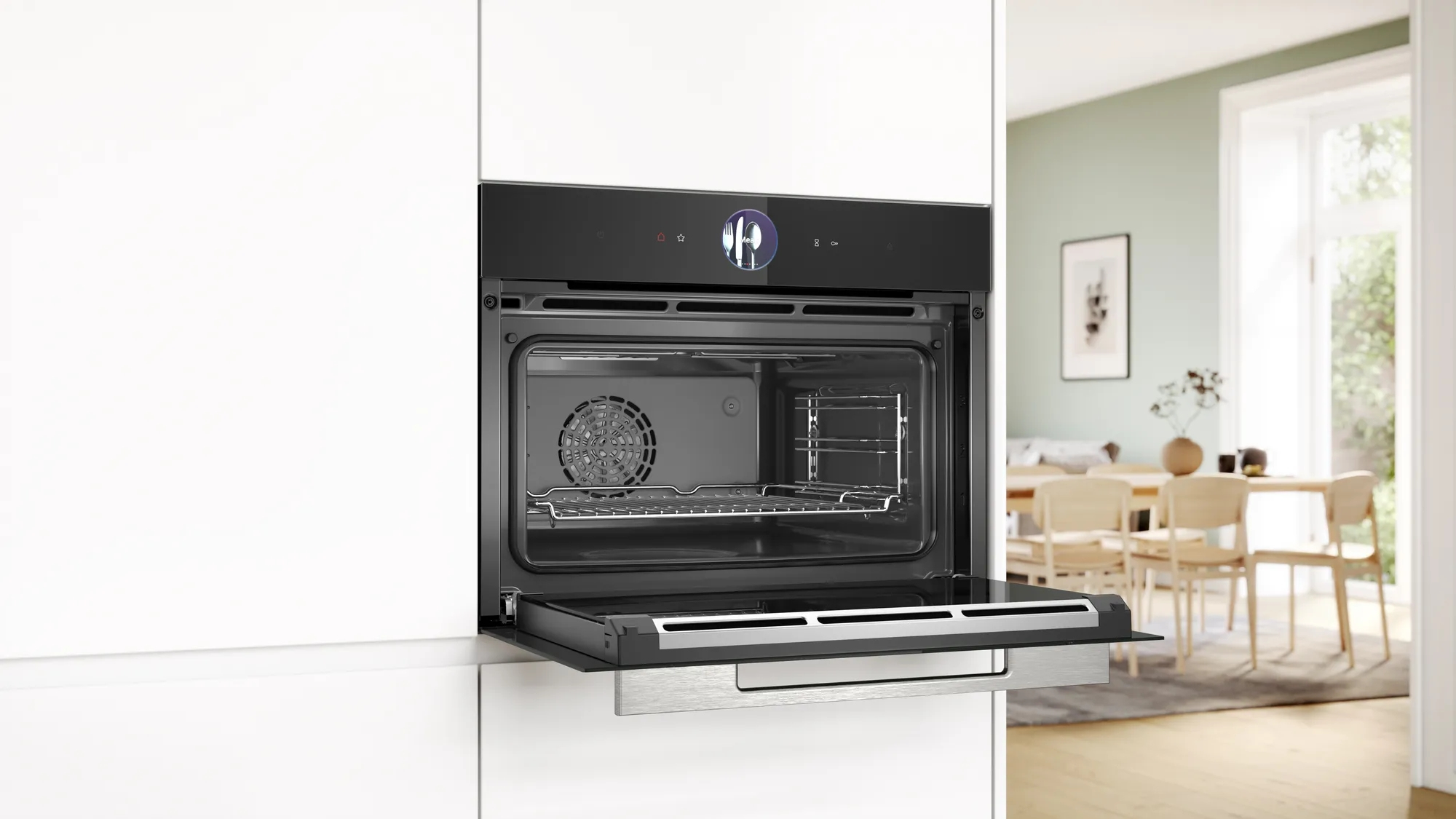 Forno incassato nero con porta aperta in una cucina bianca, con zona pranzo luminosa sullo sfondo.