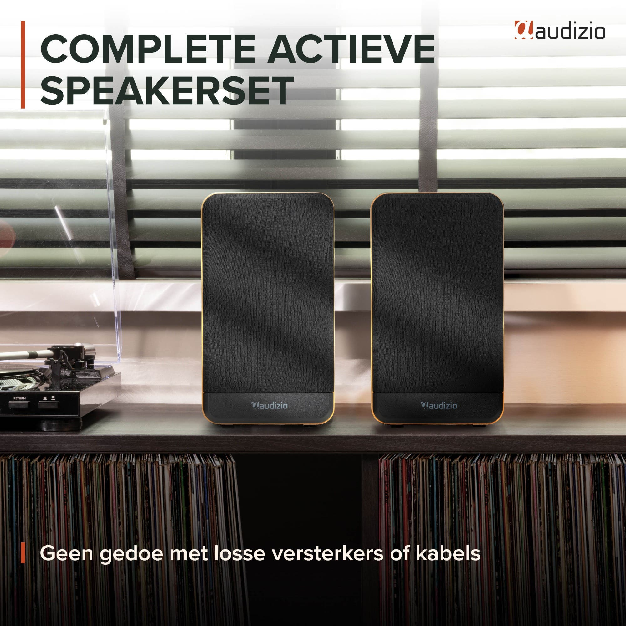 Dos altavoces negros Audizio en una mesa de madera con discos en estantes y persianas al fondo.
