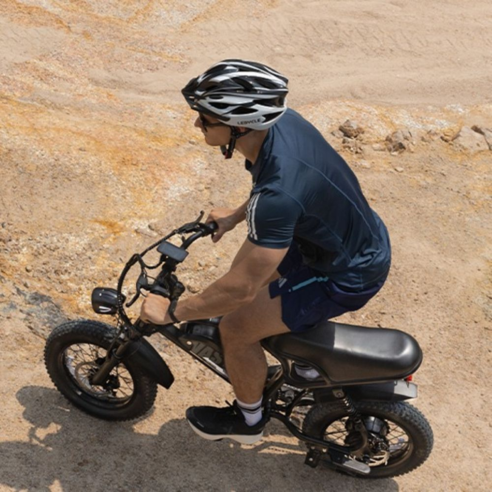Un hombre con casco conduce una pequeña bicicleta eléctrica negra en suelo arenoso.