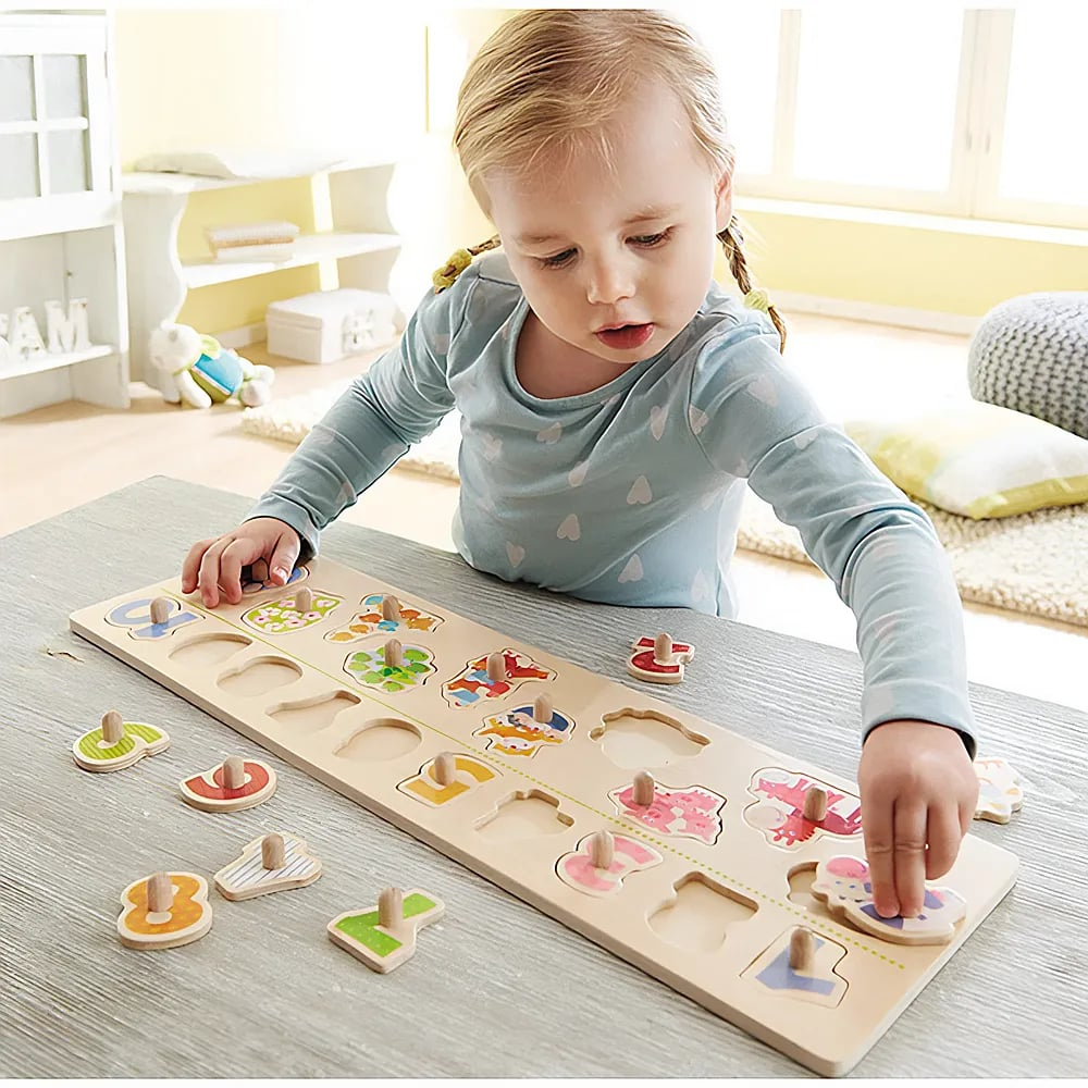 Kleinkind sitzt am Tisch und spielt mit einem Holzzahlenpuzzle in einem hellen Raum.