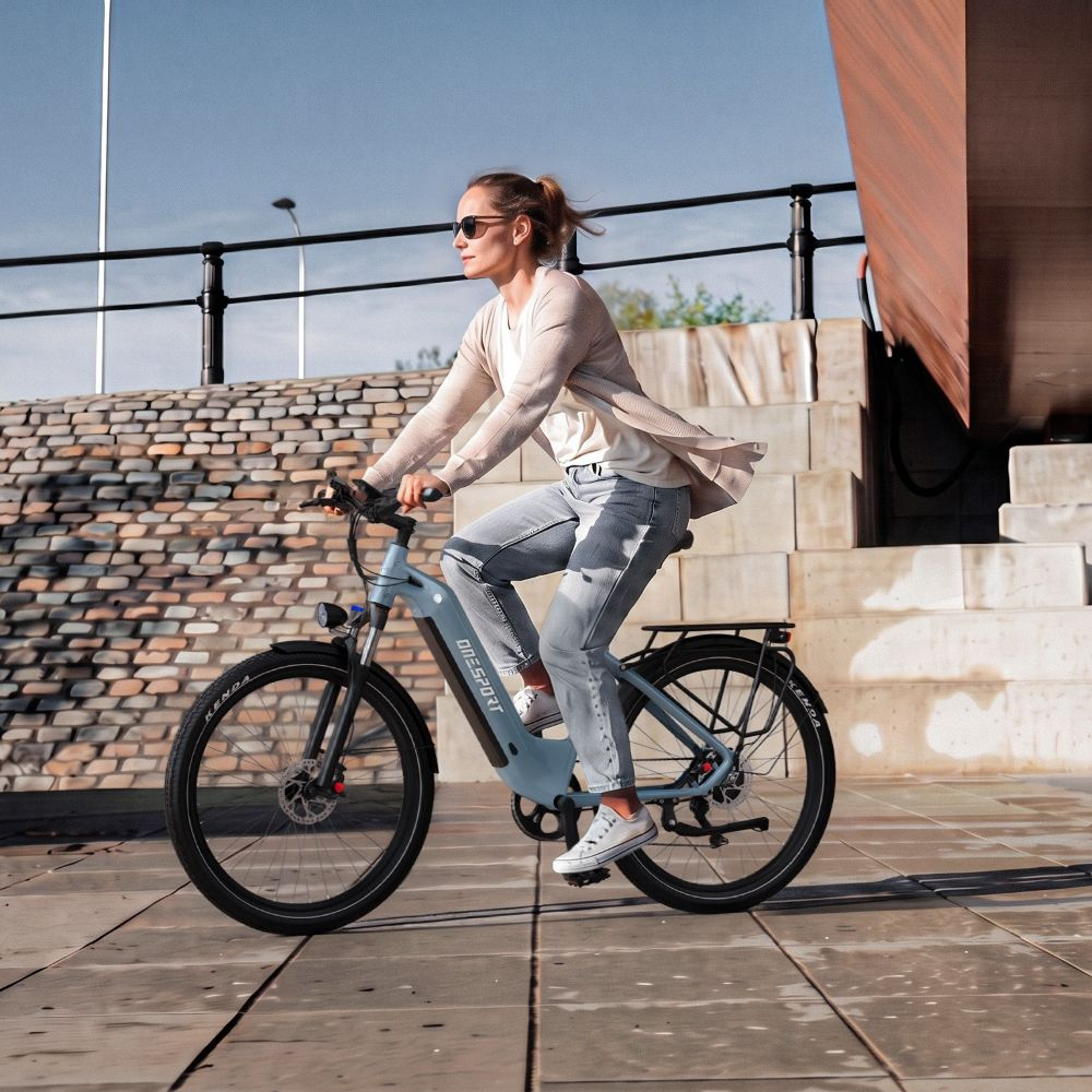 Una mujer con gafas de sol monta en una bicicleta eléctrica azul claro en una plaza soleada frente a unas escaleras.