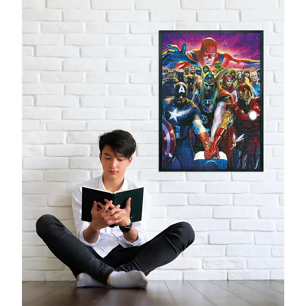 A young man sits cross-legged on the floor against a white brick wall, reading a book below a framed superhero puzzle.