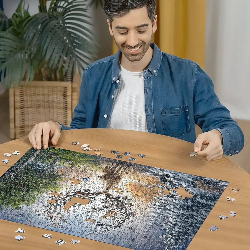 Ein lächelnder Mann in blauem Hemd sitzt an einem Holztisch und legt ein großes Puzzle mit einer Naturszene.