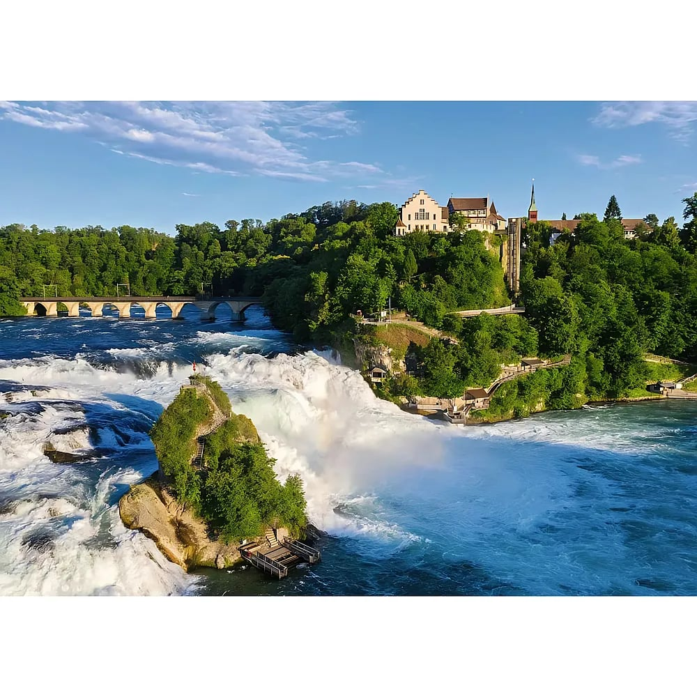 Une cascade puissante avec un îlot rocheux, un pont et des bâtiments dans la verdure sous un ciel bleu.